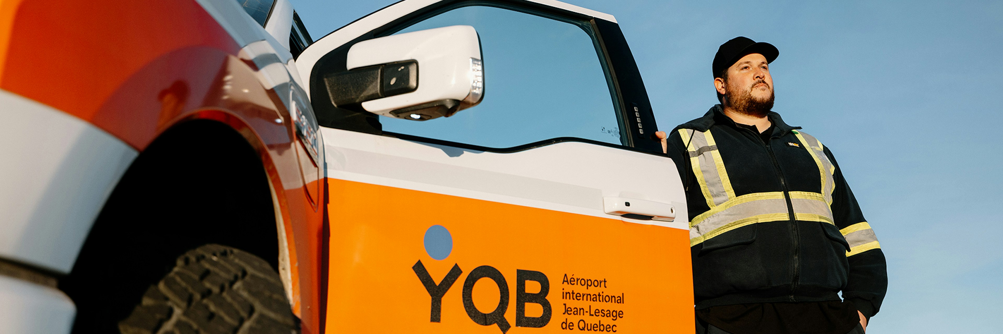 Under a clear blue sky, a man wearing a safety vest stands next to a vehicle bearing the inscription “YQB Jean-Lesage International Airport, Quebec City.”.