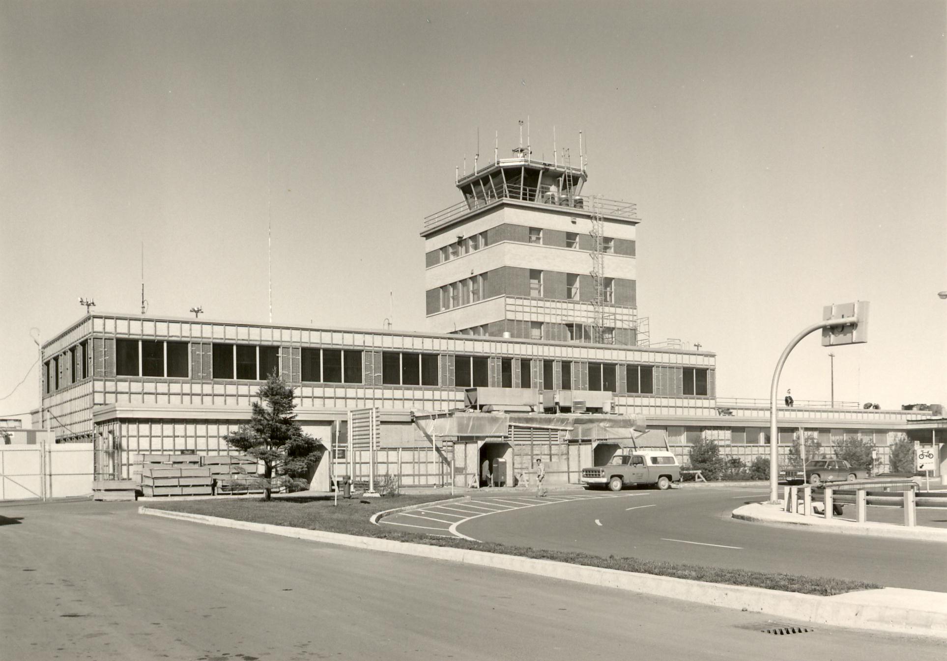 Photo en noir et blanc d'un terminal d'aéroport avec une tour de contrôle, des véhicules garés et un aménagement paysager minimal sur une allée incurvée située devant.