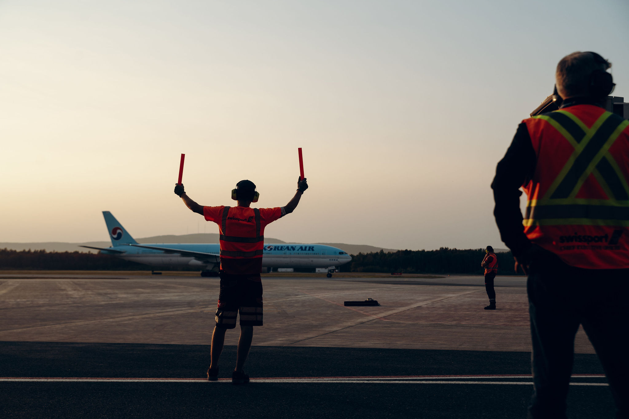 Un membre du personnel au sol, vêtu d'un gilet de sécurité, guide à l'aide de bâtons un avion de Korean Air sur la piste au coucher du soleil, tandis que d'autres membres du personnel se tiennent à proximité.