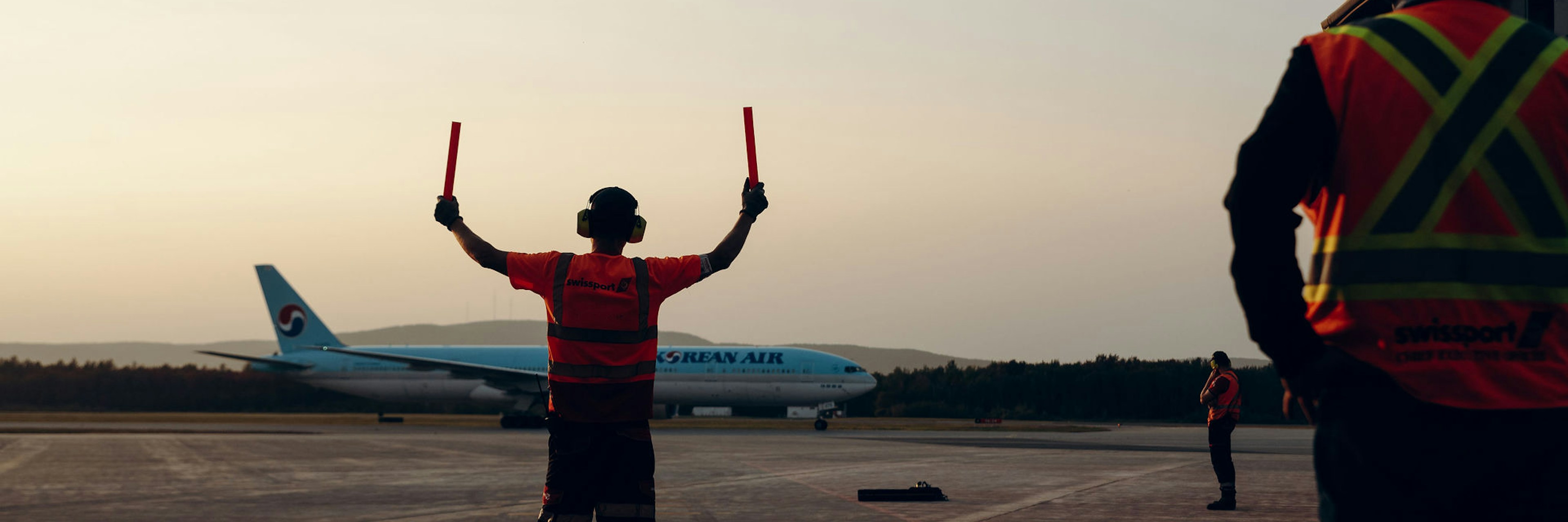 Un membre du personnel au sol, vêtu d'un gilet de sécurité, guide à l'aide de bâtons un avion de Korean Air sur la piste au coucher du soleil, tandis que d'autres membres du personnel se tiennent à proximité.