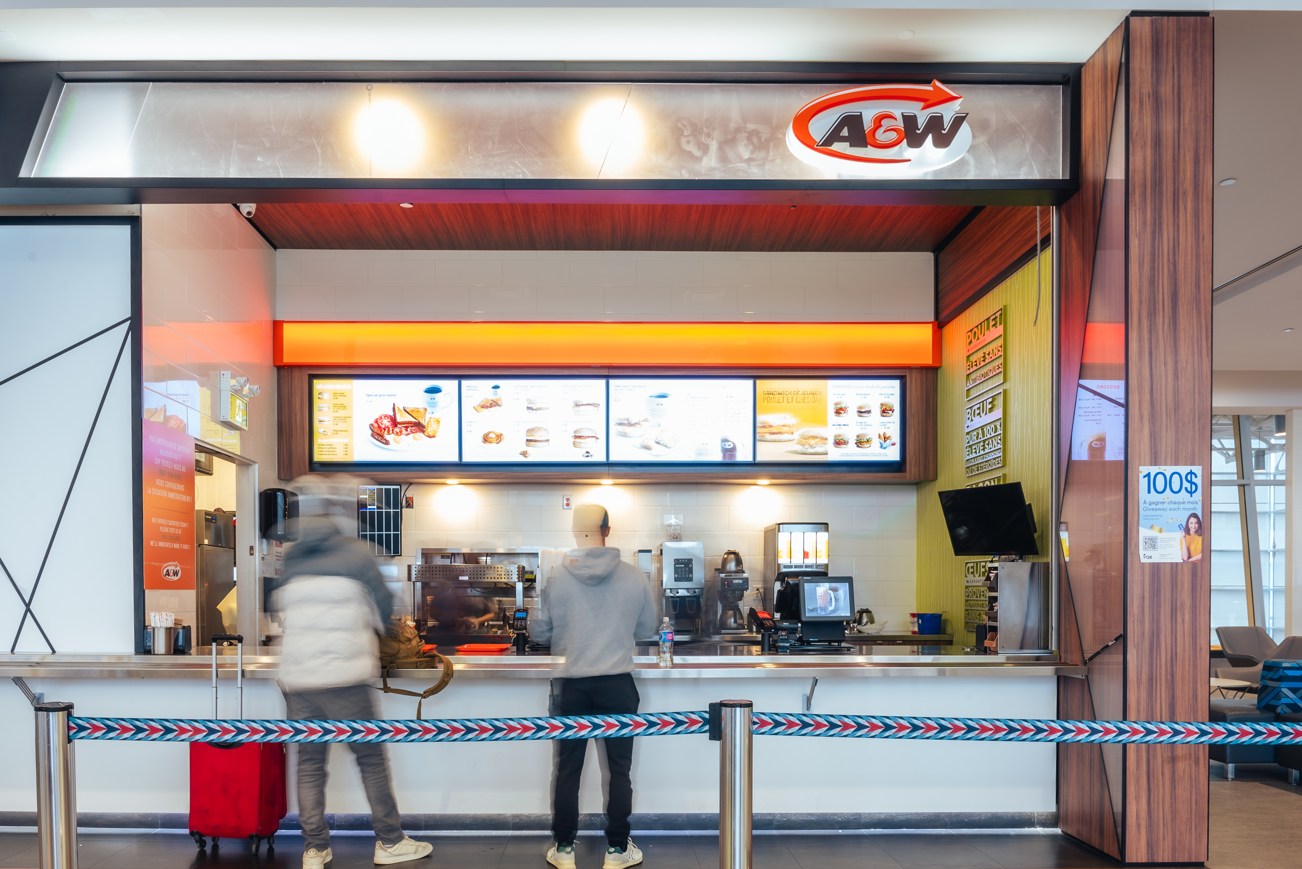 Deux personnes se tiennent au comptoir d'un restaurant de restauration rapide A&W, les yeux rivés sur le menu affiché au-dessus. Le comptoir est moderne, et une barrière en corde sépare la zone de commande du reste de l'espace.