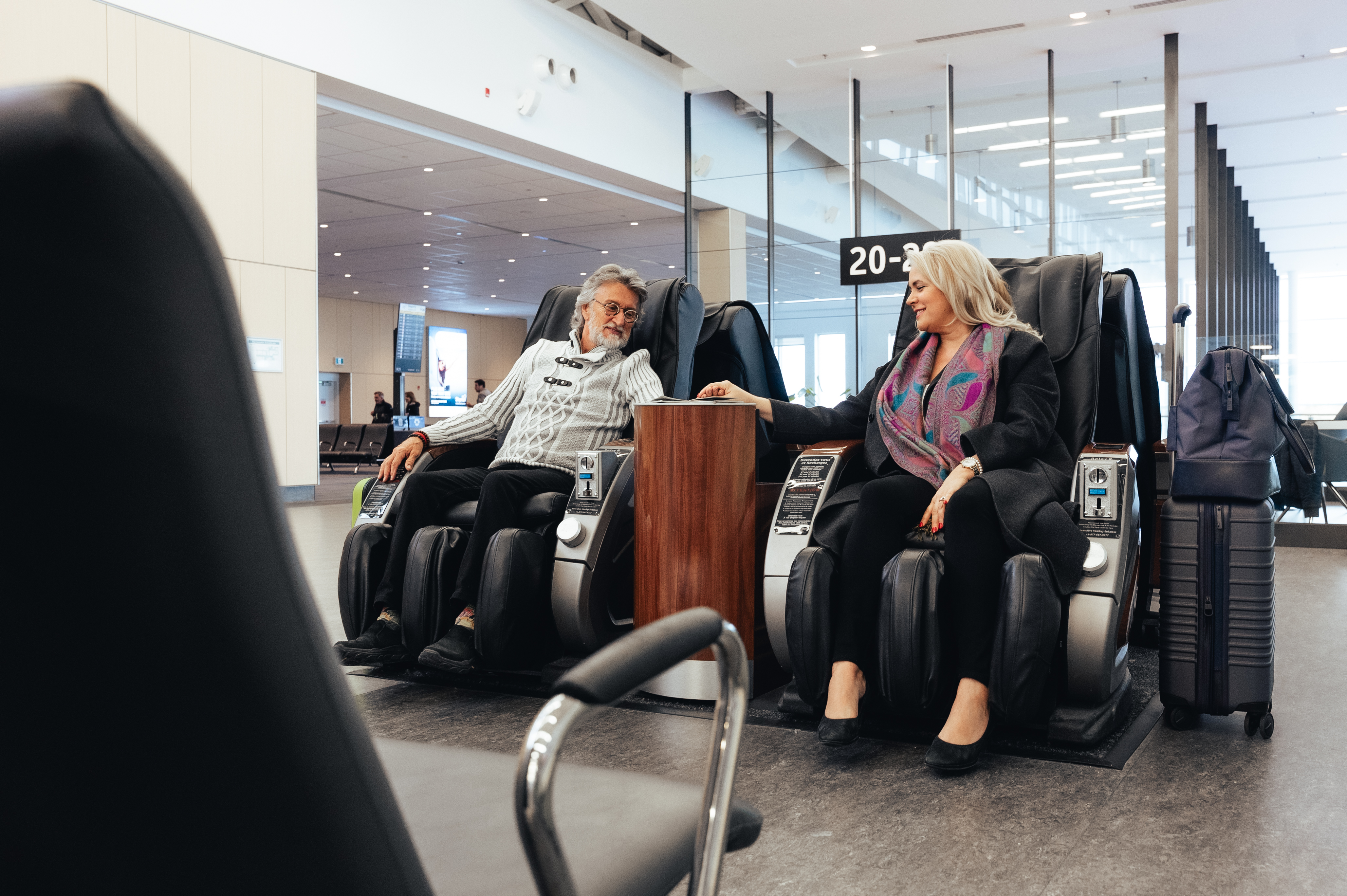 Un couple d'âge mûr est assis dans des fauteuils de massage dans la zone d'embarquement d'un aéroport ; ils se sourient et se tiennent la main, leurs bagages à proximité.