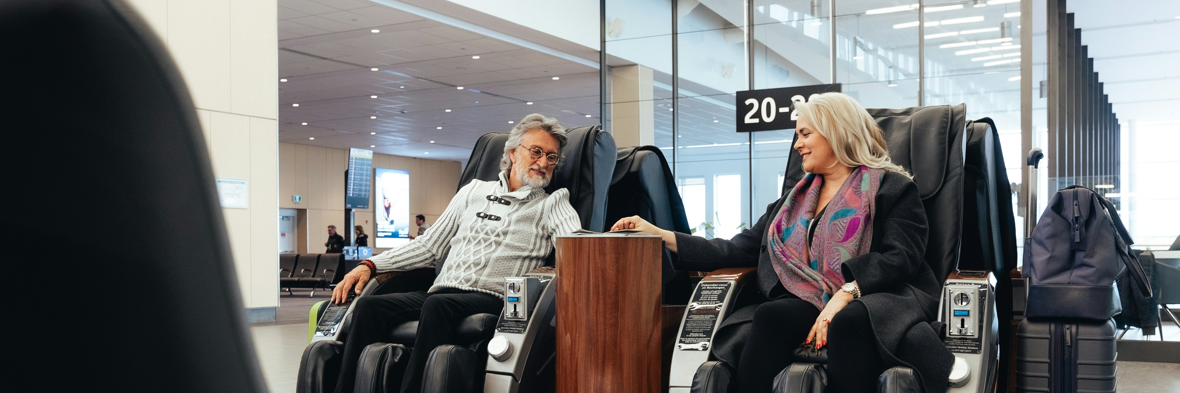 Un couple d'âge mûr est assis dans des fauteuils de massage dans la zone d'embarquement d'un aéroport ; ils se sourient et se tiennent la main, leurs bagages à proximité.