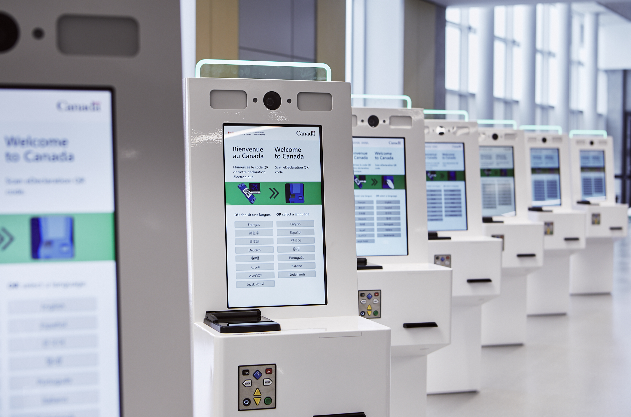 Une rangée de bornes électroniques en libre-service, dont les écrans affichent le message « Bienvenue au Canada » en anglais et en français, dans un aéroport moderne et bien éclairé ou dans une zone de contrôle aux frontières.