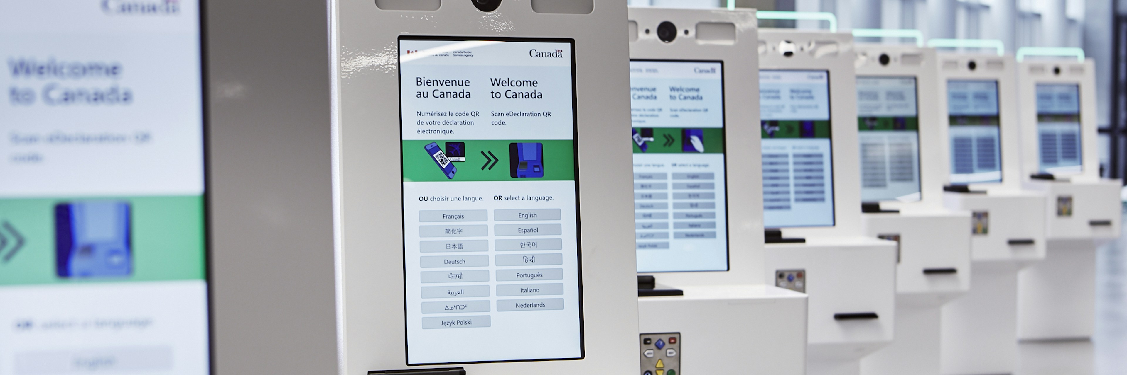 Une rangée de bornes électroniques en libre-service, dont les écrans affichent le message « Bienvenue au Canada » en anglais et en français, dans un aéroport moderne et bien éclairé ou dans une zone de contrôle aux frontières.