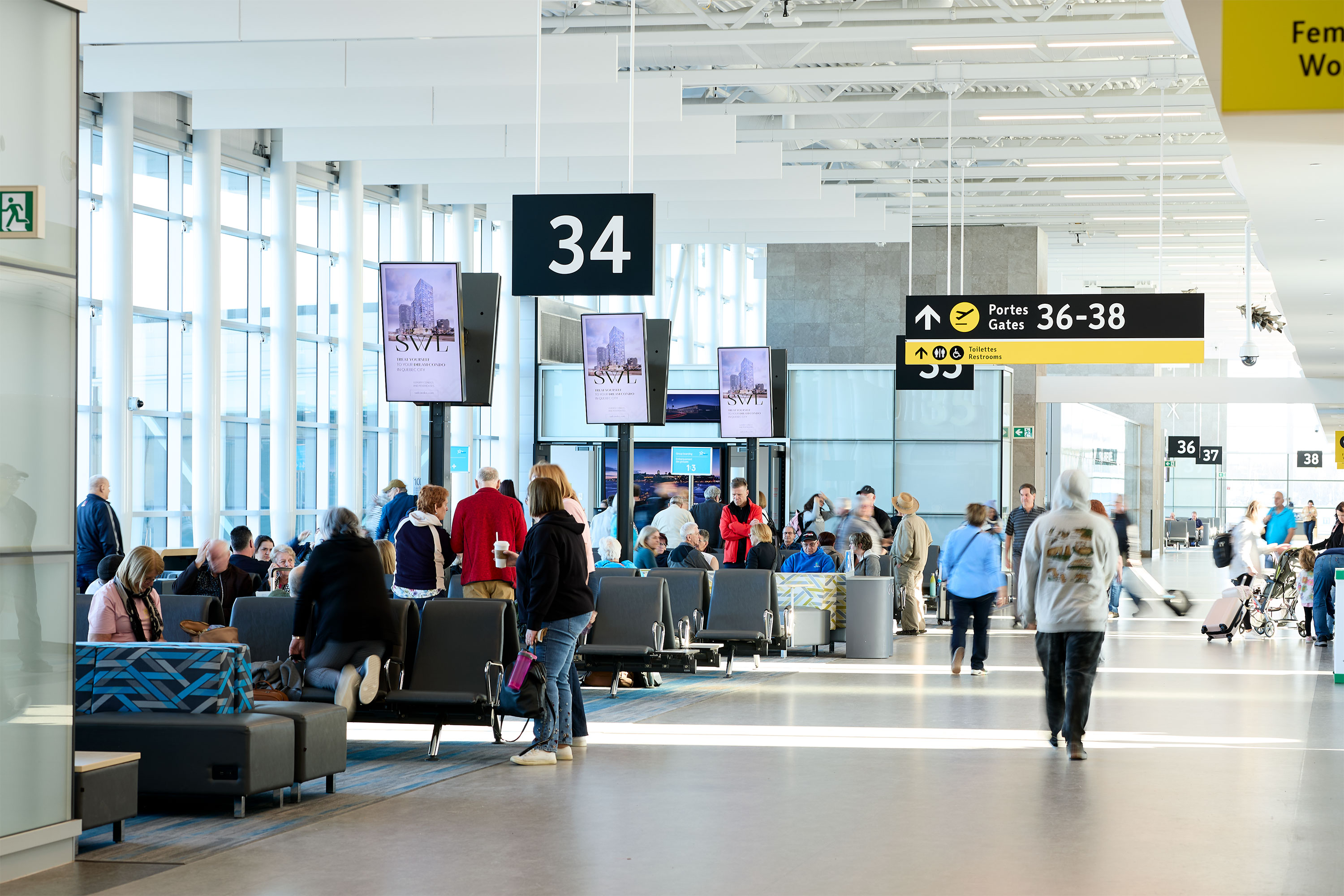 Les passagers attendent et circulent près de la porte 34 dans un terminal d'aéroport lumineux et moderne, doté de grandes baies vitrées, d'espaces pour s'asseoir et de panneaux indicateurs vers les portes 36 à 38, visibles à l'arrière-plan.
