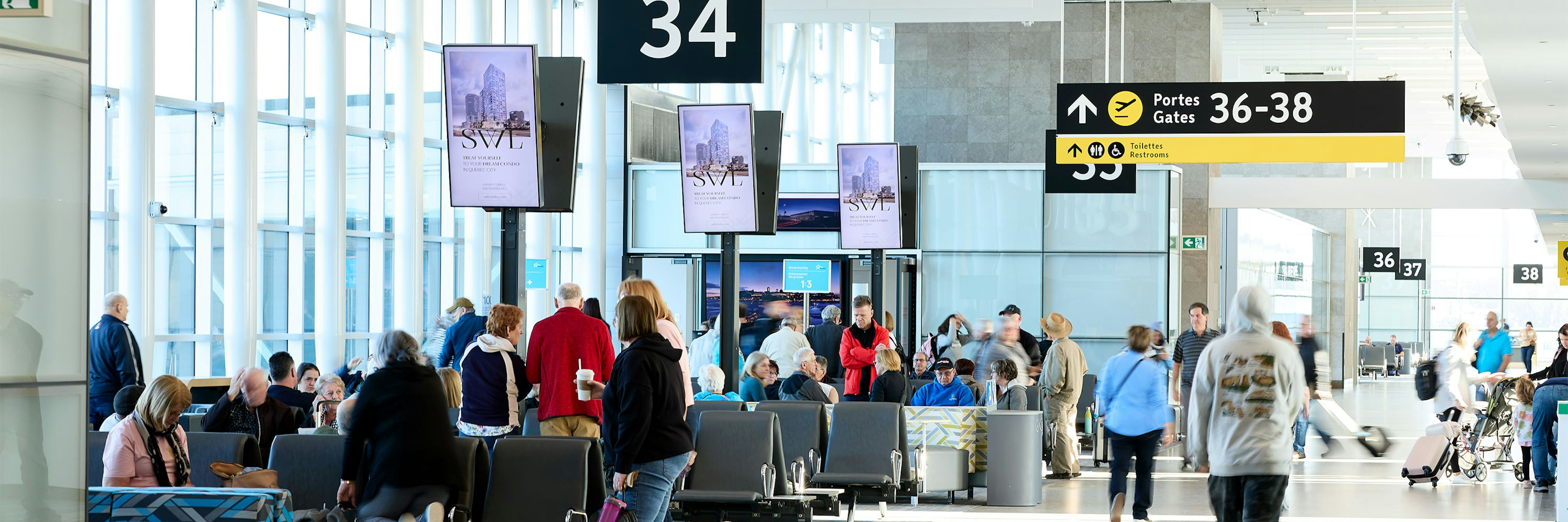 Les passagers attendent et circulent près de la porte 34 dans un terminal d'aéroport lumineux et moderne, doté de grandes baies vitrées, d'espaces pour s'asseoir et de panneaux indicateurs vers les portes 36 à 38, visibles à l'arrière-plan.