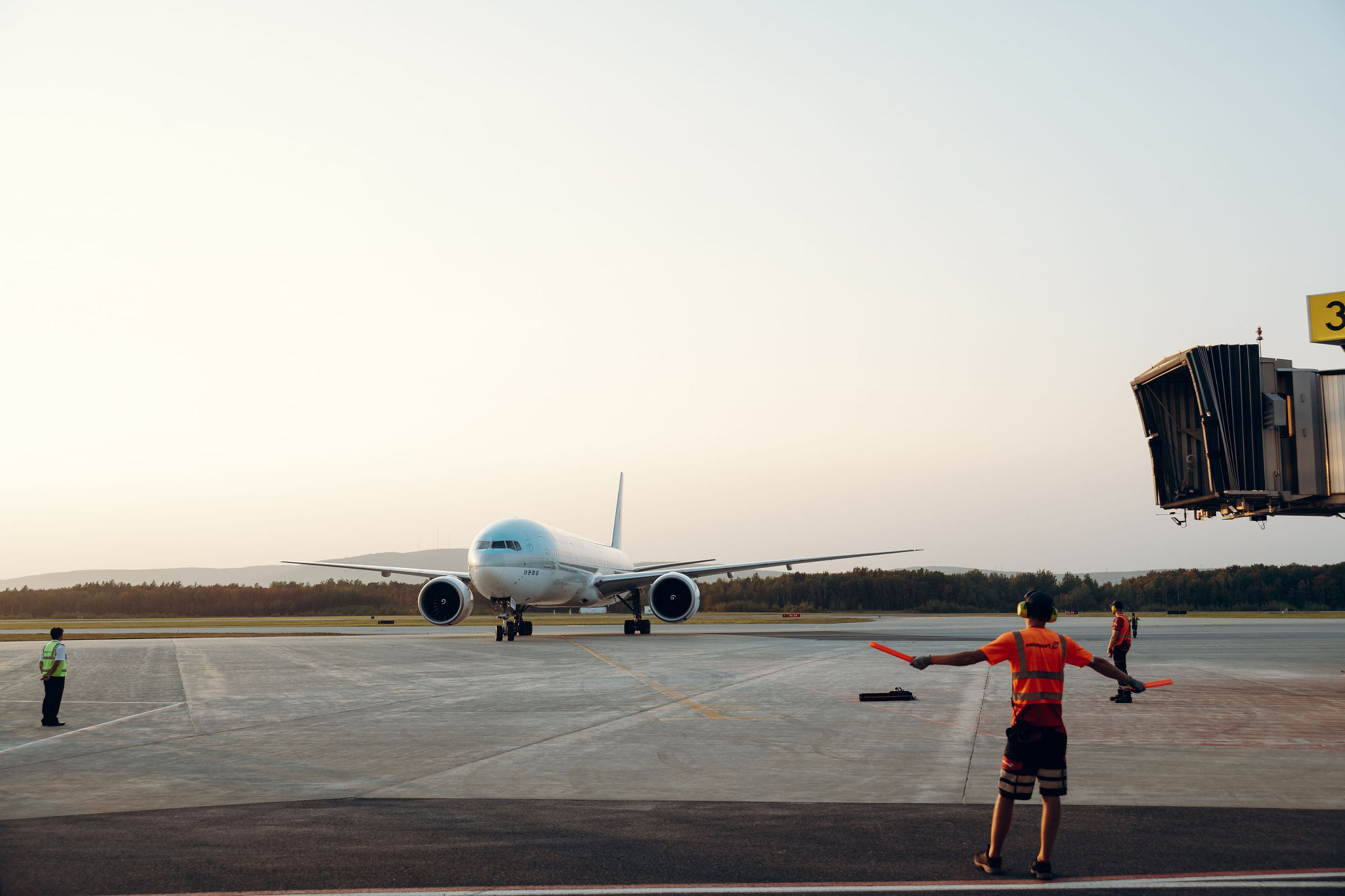 Un avion à réaction roule sur la piste en direction d'une porte d'embarquement, tandis que trois membres du personnel au sol le guident à l'aide de bâtons dans la lumière du début de soirée.