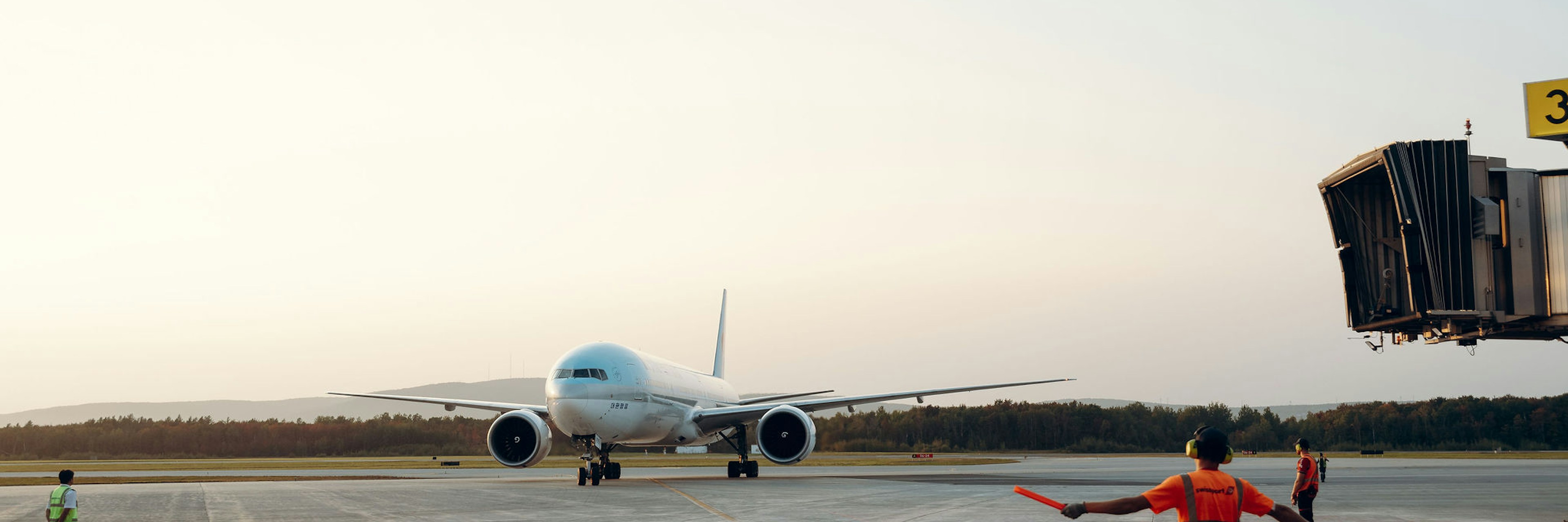 Un avion à réaction roule sur la piste en direction d'une porte d'embarquement, tandis que trois membres du personnel au sol le guident à l'aide de bâtons dans la lumière du début de soirée.