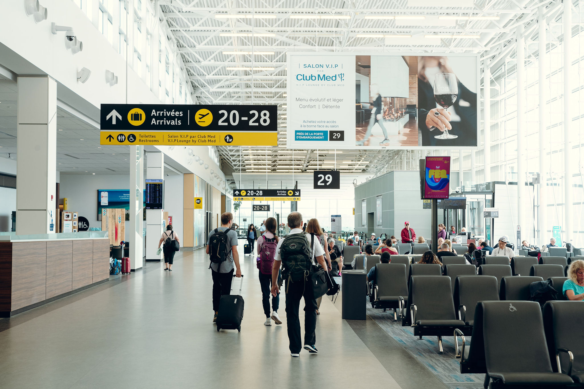 Des voyageurs accompagnés de leurs bagages traversent un terminal d'aéroport lumineux et moderne, doté de hauts plafonds, de grandes baies vitrées, de panneaux indiquant les portes d'embarquement et de personnes assises dans les zones d'attente.