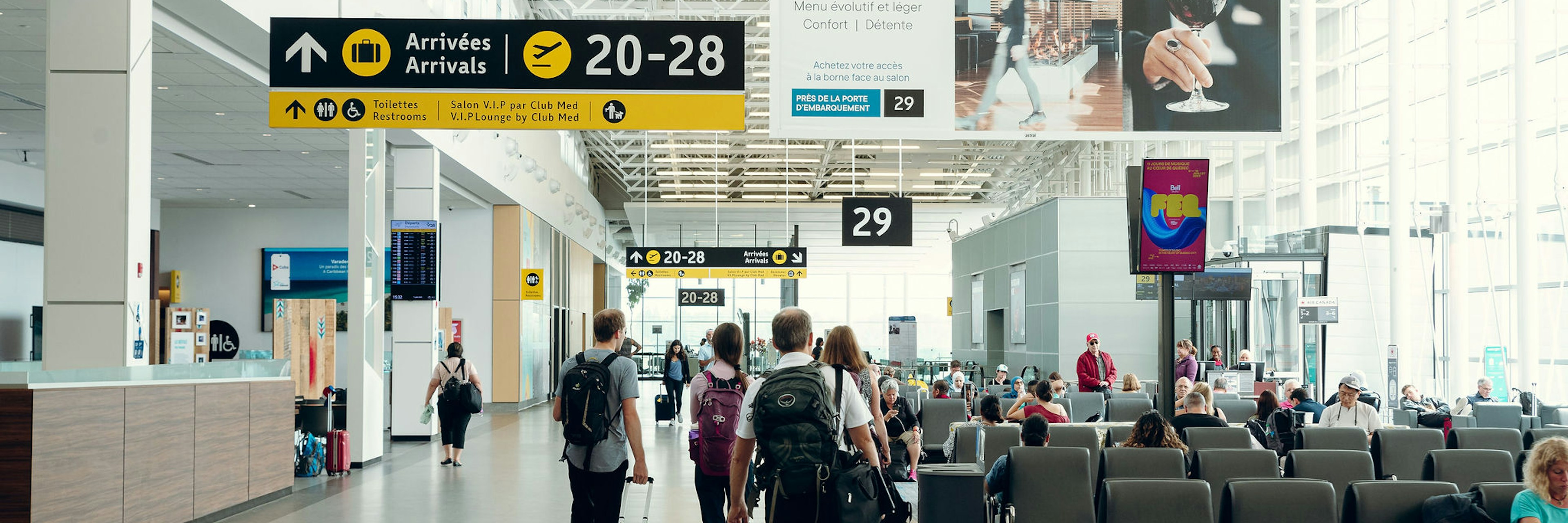 Des voyageurs accompagnés de leurs bagages traversent un terminal d'aéroport lumineux et moderne, doté de hauts plafonds, de grandes baies vitrées, de panneaux indiquant les portes d'embarquement et de personnes assises dans les zones d'attente.
