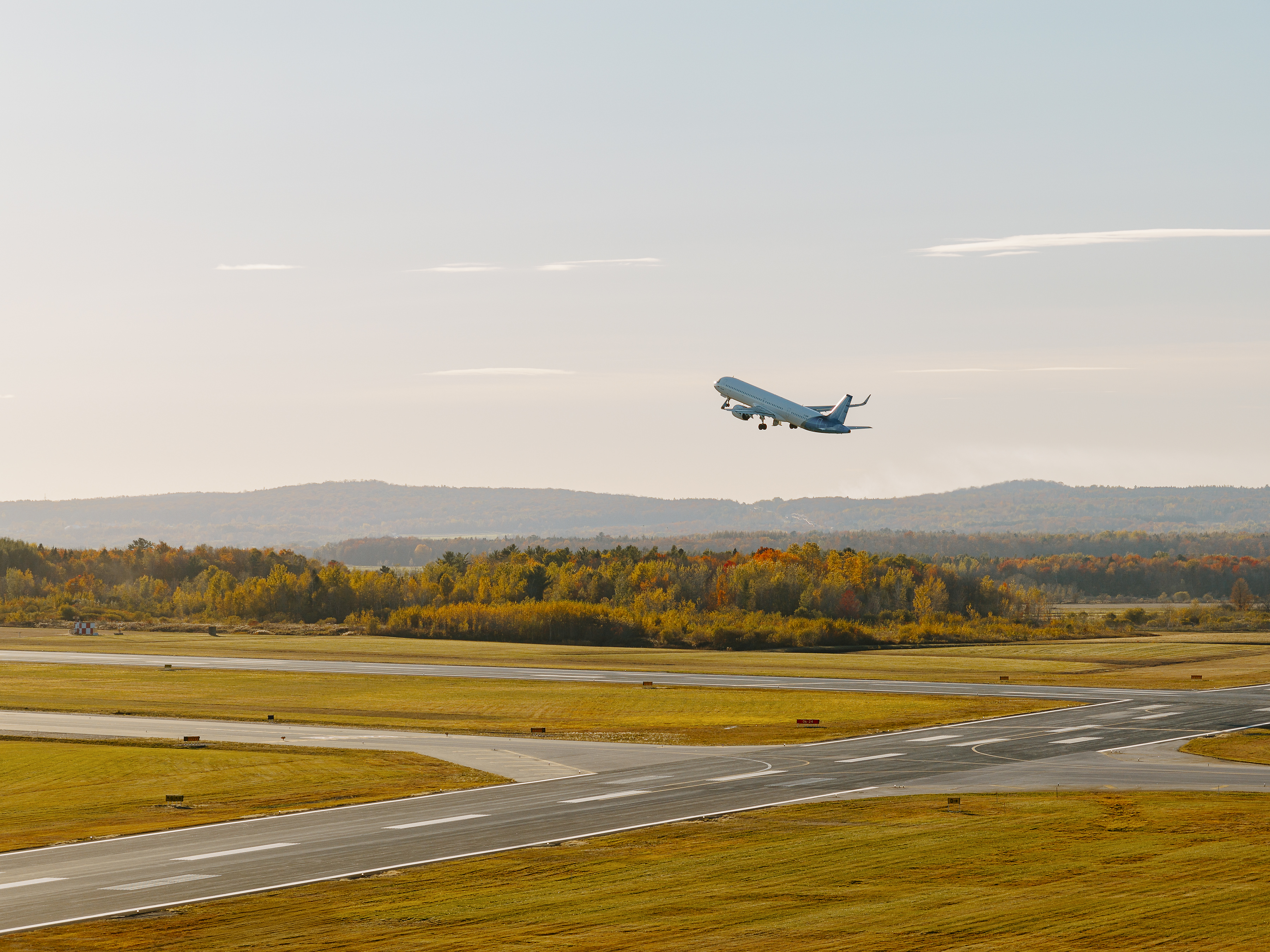 Un avion de ligne décolle d'une piste et s'élève dans les airs au-dessus d'un paysage automnal, où les arbres arborent leurs couleurs d'automne sous un ciel dégagé.