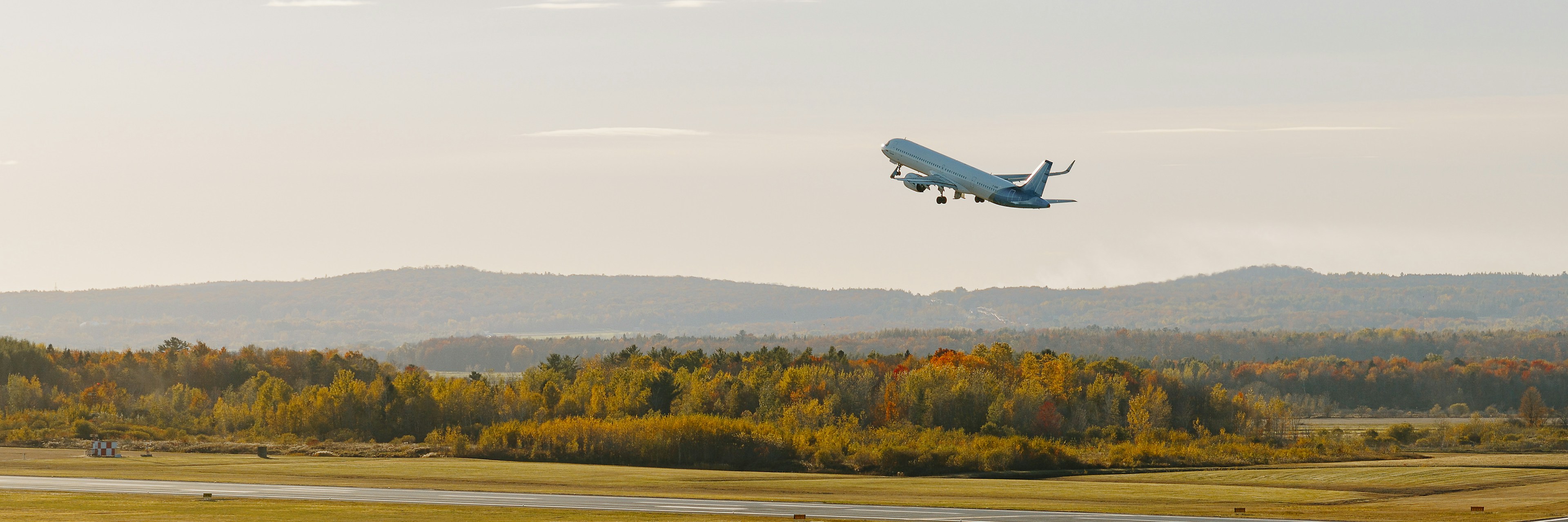 Un avion de ligne décolle d'une piste et s'élève dans les airs au-dessus d'un paysage automnal, où les arbres arborent leurs couleurs d'automne sous un ciel dégagé.