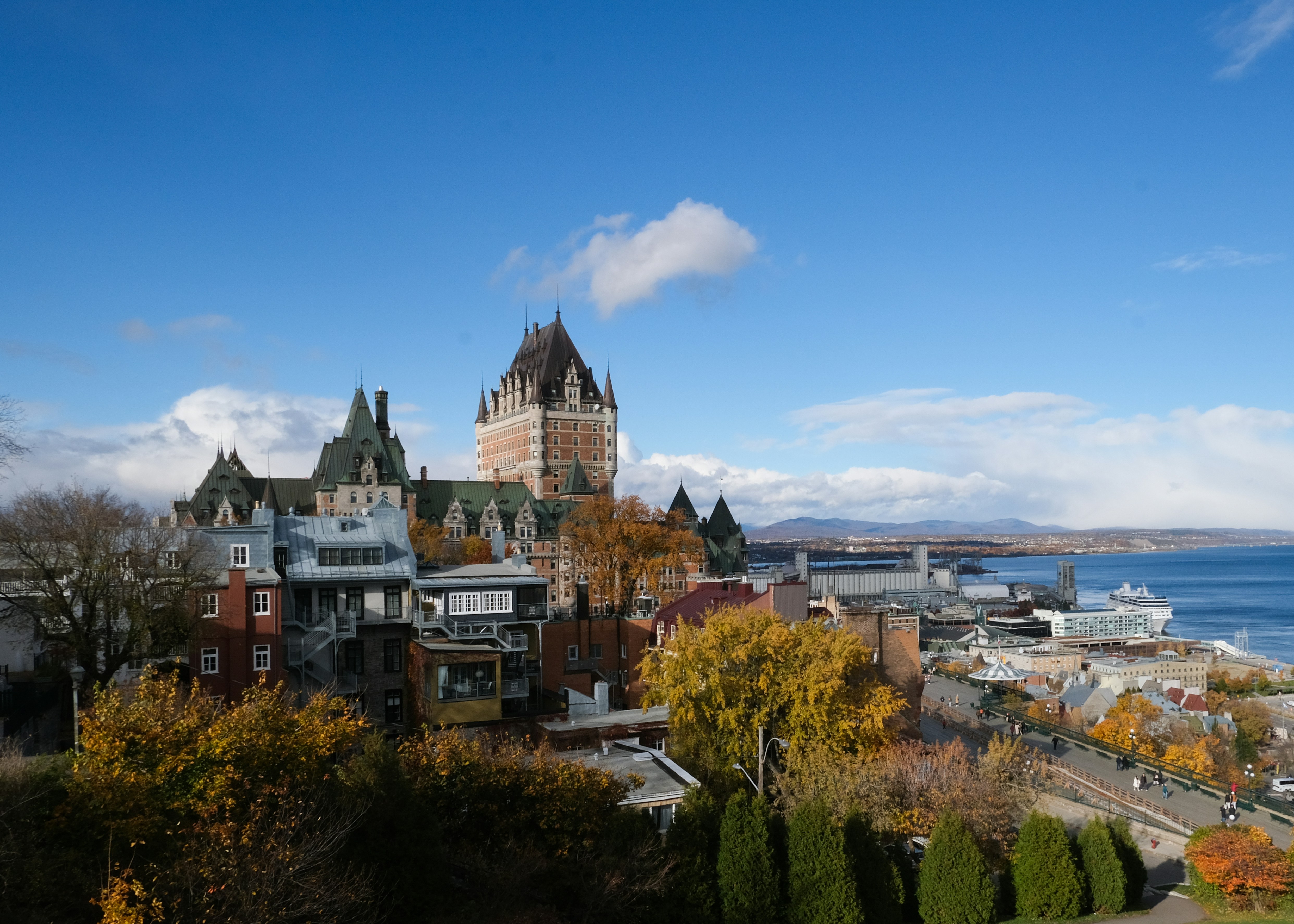 Vue du Château Frontenac entouré de feuillages d'automne, avec des bâtiments historiques, une rivière et des montagnes au loin, sous un ciel bleu parsemé de nuages, à Québec.