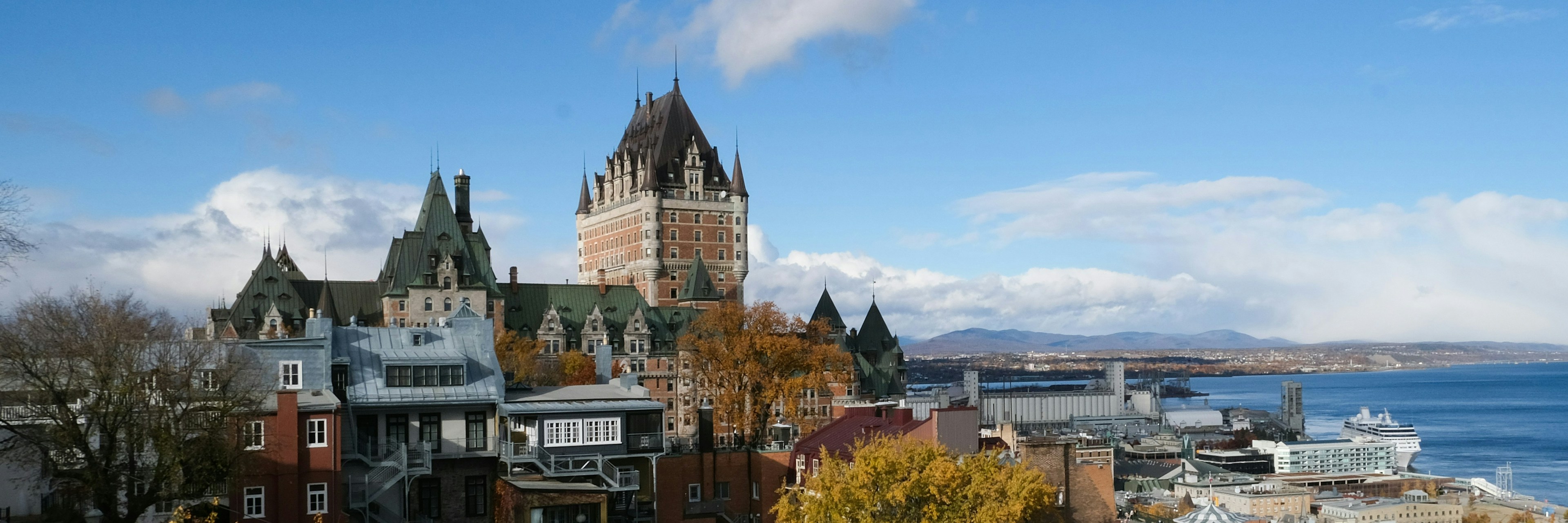 Vue du Château Frontenac entouré de feuillages d'automne, avec des bâtiments historiques, une rivière et des montagnes au loin, sous un ciel bleu parsemé de nuages, à Québec.