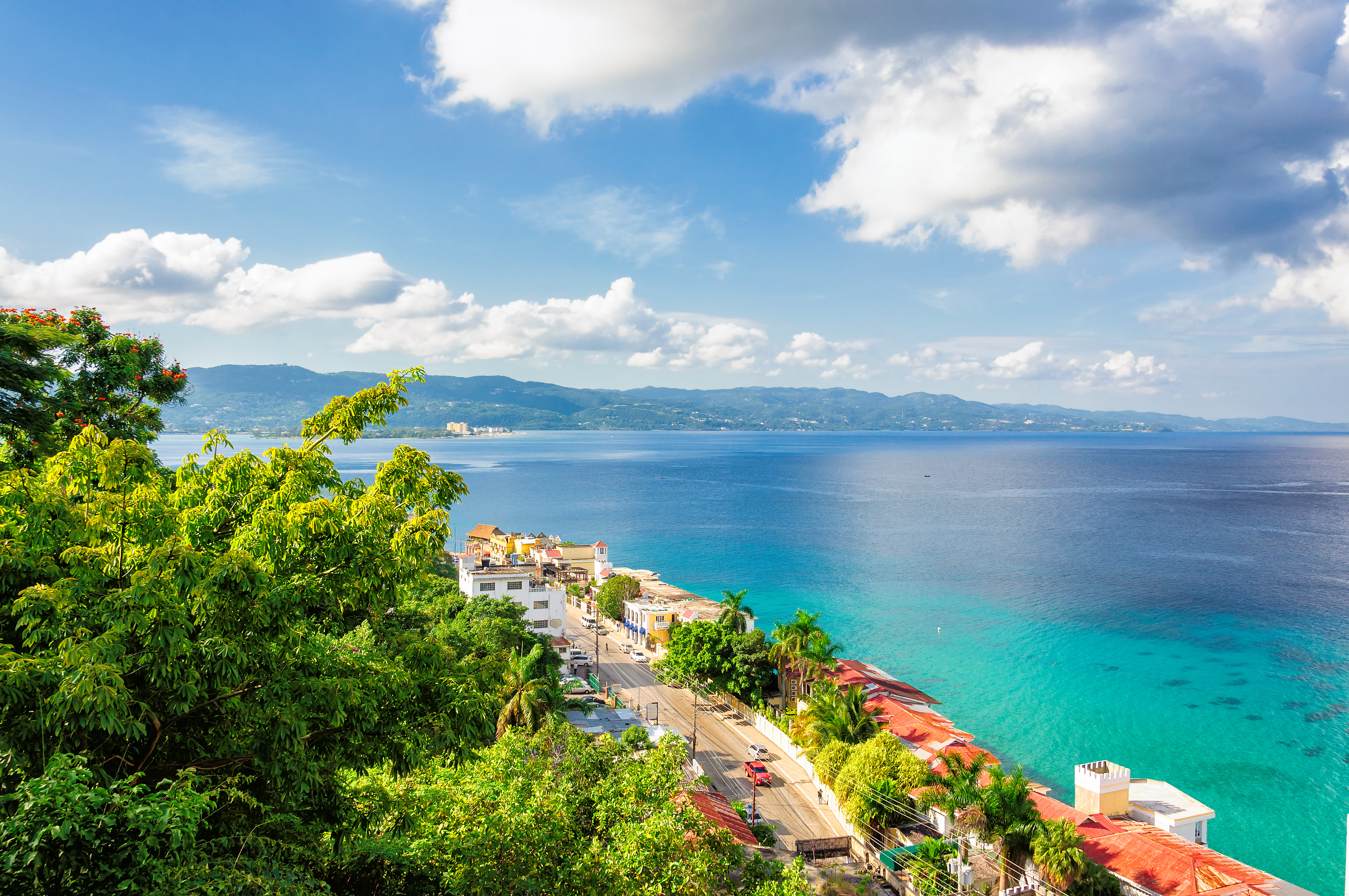 A scenic view of a coastal town with lush green trees, turquoise-blue ocean water, rooftops, and distant mountains under a partly cloudy sky.