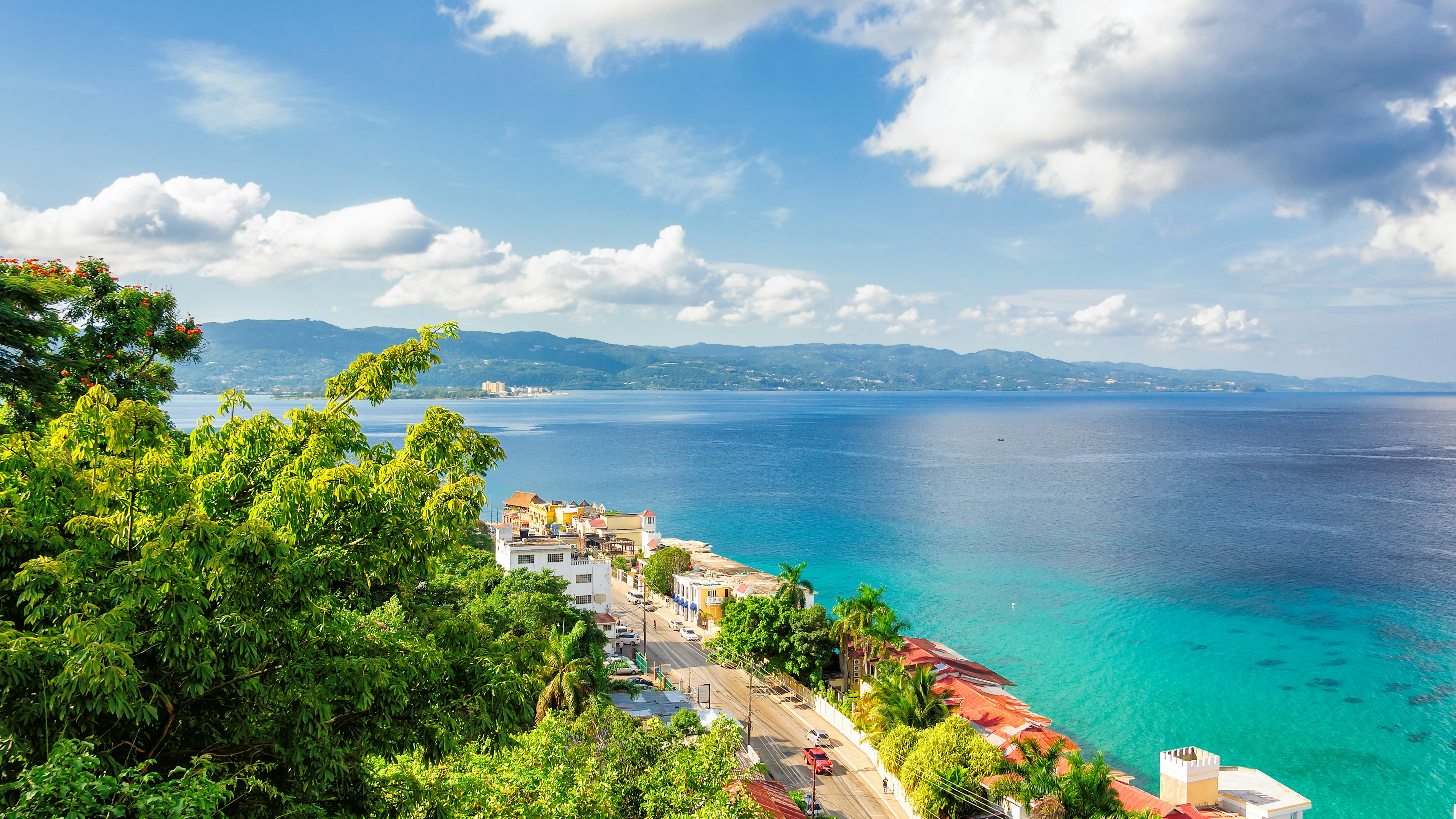A scenic view of a coastal town with lush green trees, turquoise-blue ocean water, rooftops, and distant mountains under a partly cloudy sky.