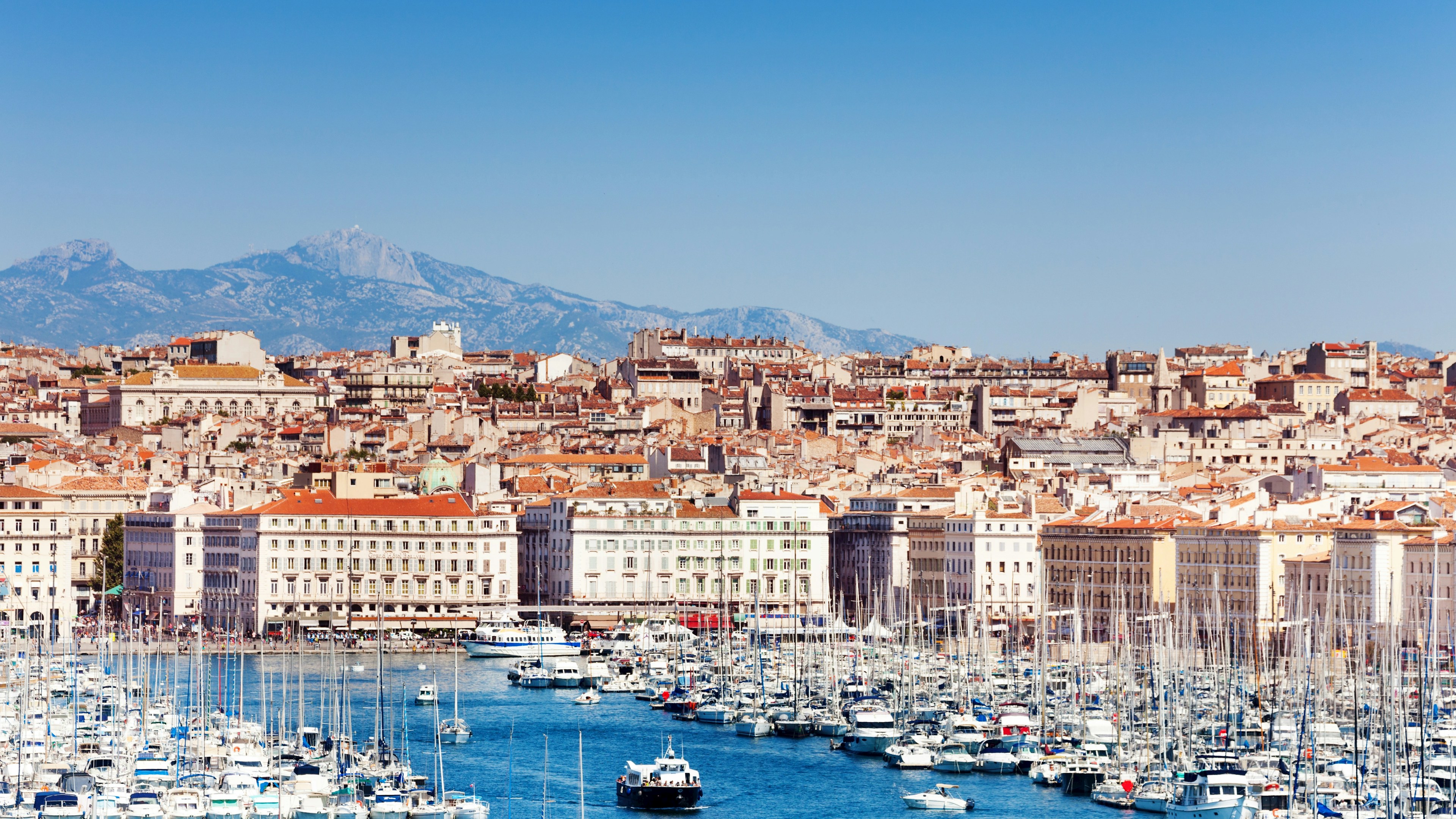 Un port de plaisance regorgeant de bateaux, face à une ville aux immeubles serrés les uns contre les autres, sous un ciel bleu limpide, avec les montagnes en arrière-plan.