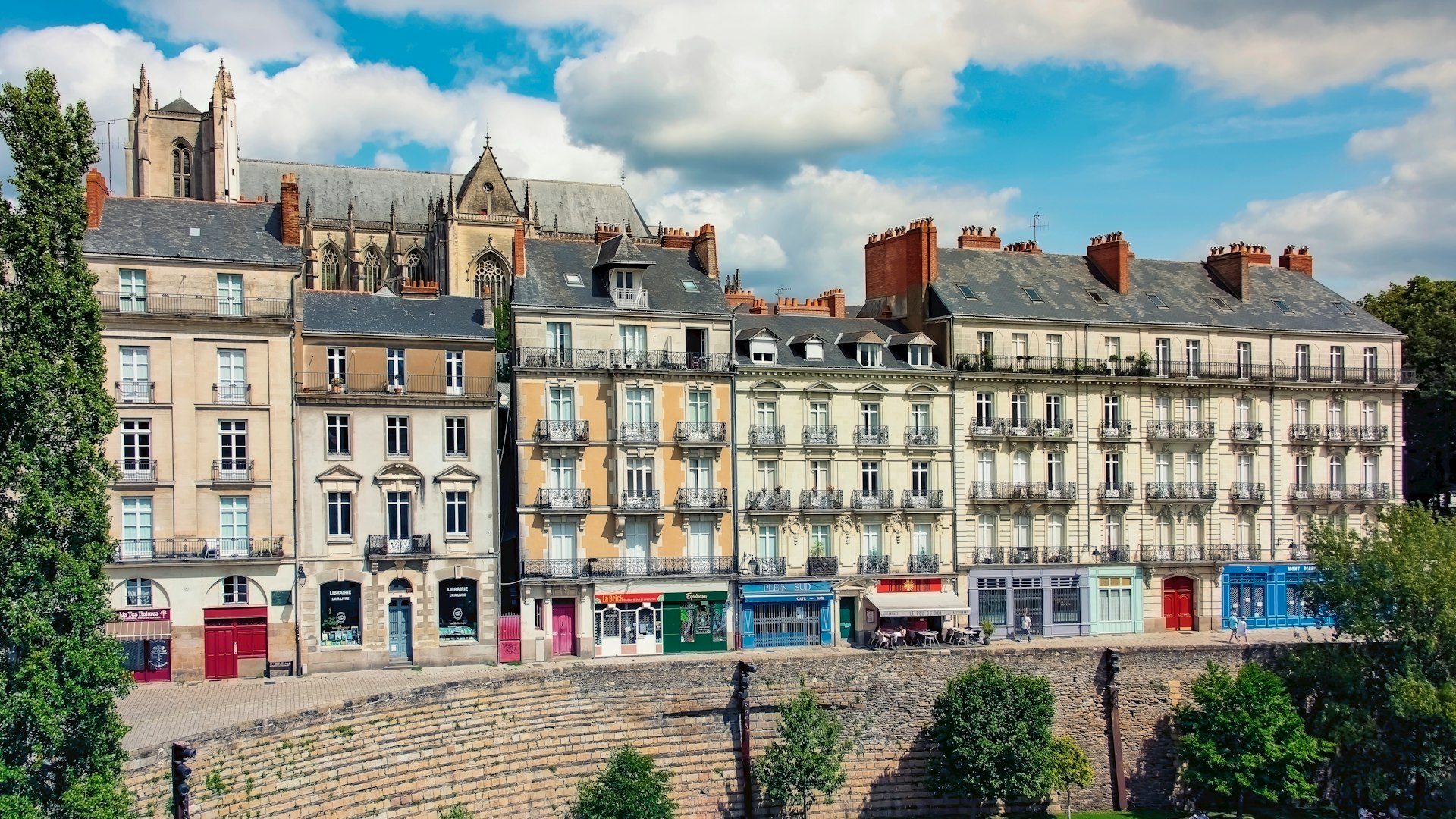 Rows of historic buildings with shops at street level stand behind a stone wall and trees under a partly cloudy sky in a European city.