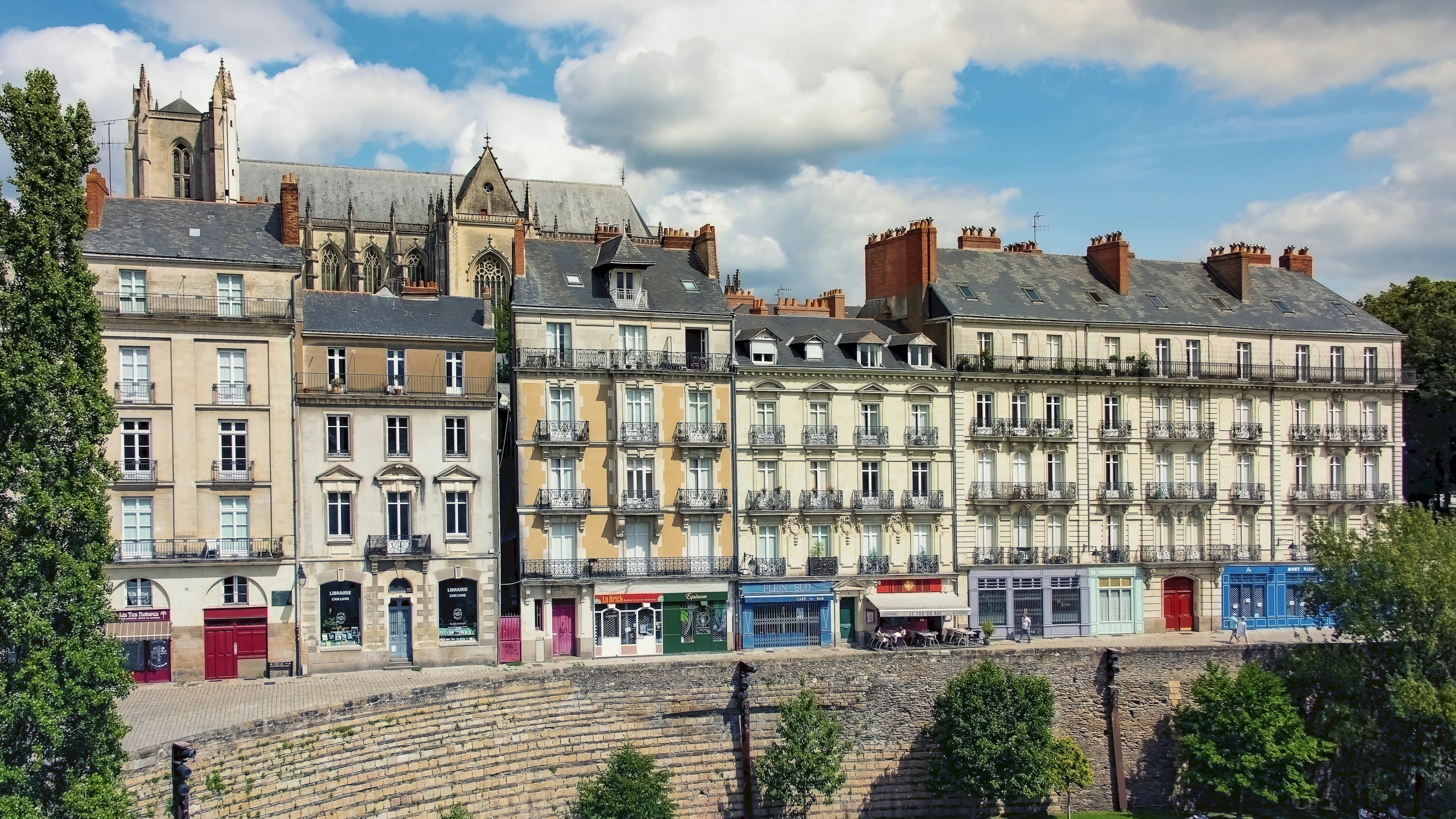 Dans une ville européenne, sous un ciel partiellement nuageux, des rangées de bâtiments historiques abritant des boutiques au rez-de-chaussée se dressent derrière un mur de pierre et des arbres.