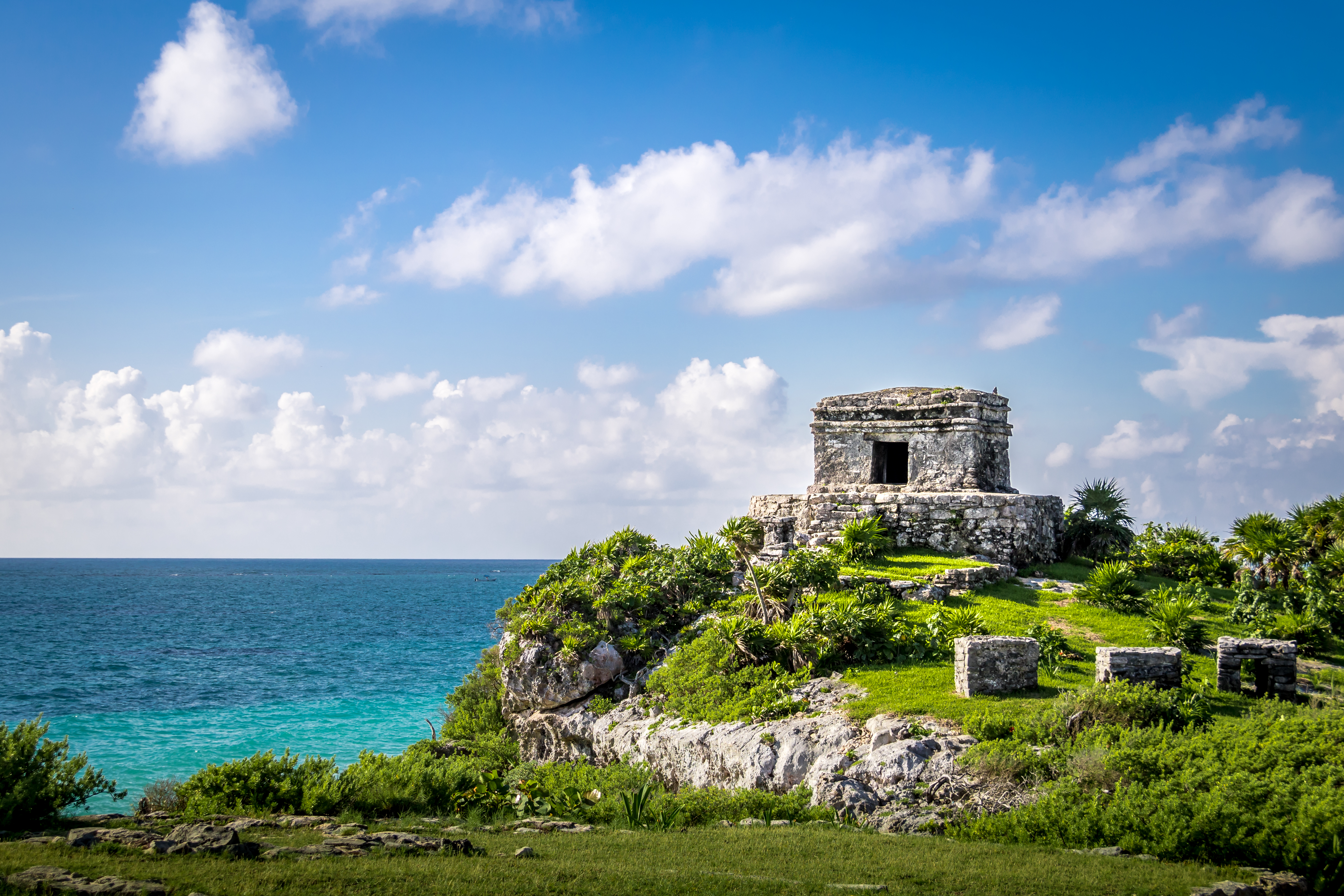 D'anciennes ruines mayas en pierre se dressent au sommet d'une falaise verdoyante et luxuriante, surplombant les eaux turquoise de l'océan, sous un ciel d'un bleu éclatant parsemé de nuages épars.