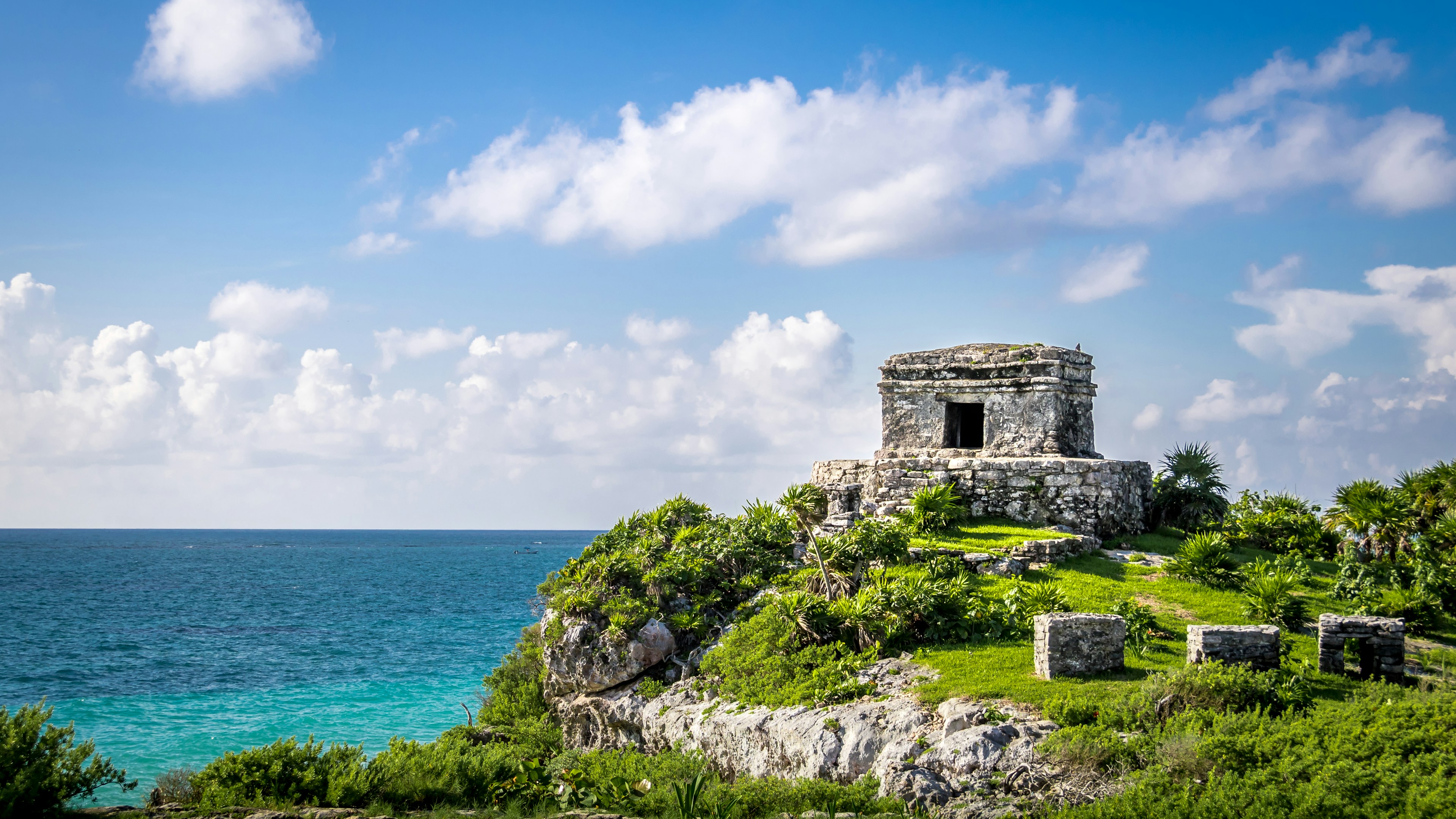 Ancient stone Mayan temple sits on a rocky cliff overlooking turquoise ocean water, surrounded by lush green plants under a bright blue sky with scattered clouds.