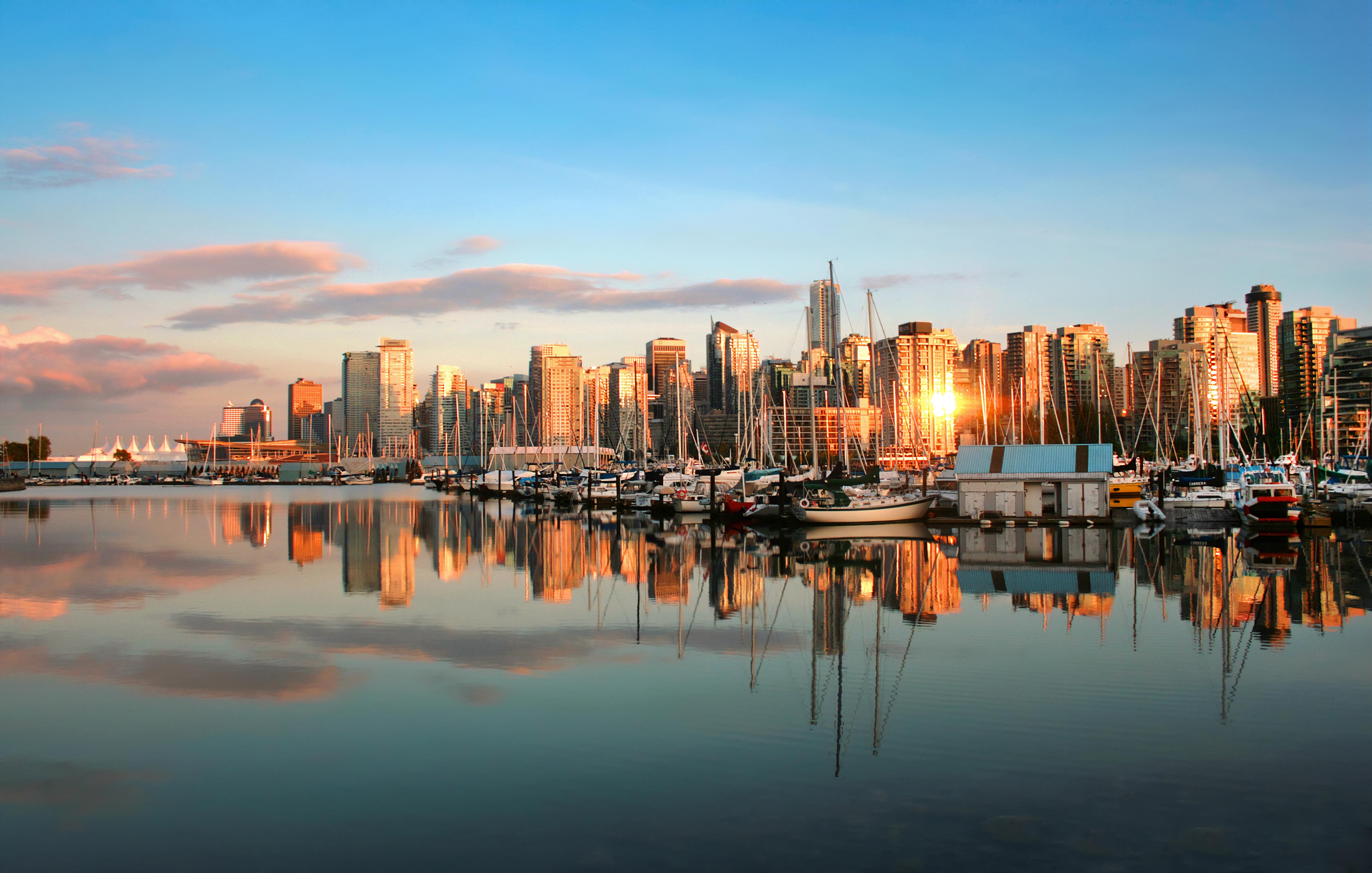 A marina where boats are moored in calm waters that reflect the city skyline at sunset, with tall buildings and clouds in the background.