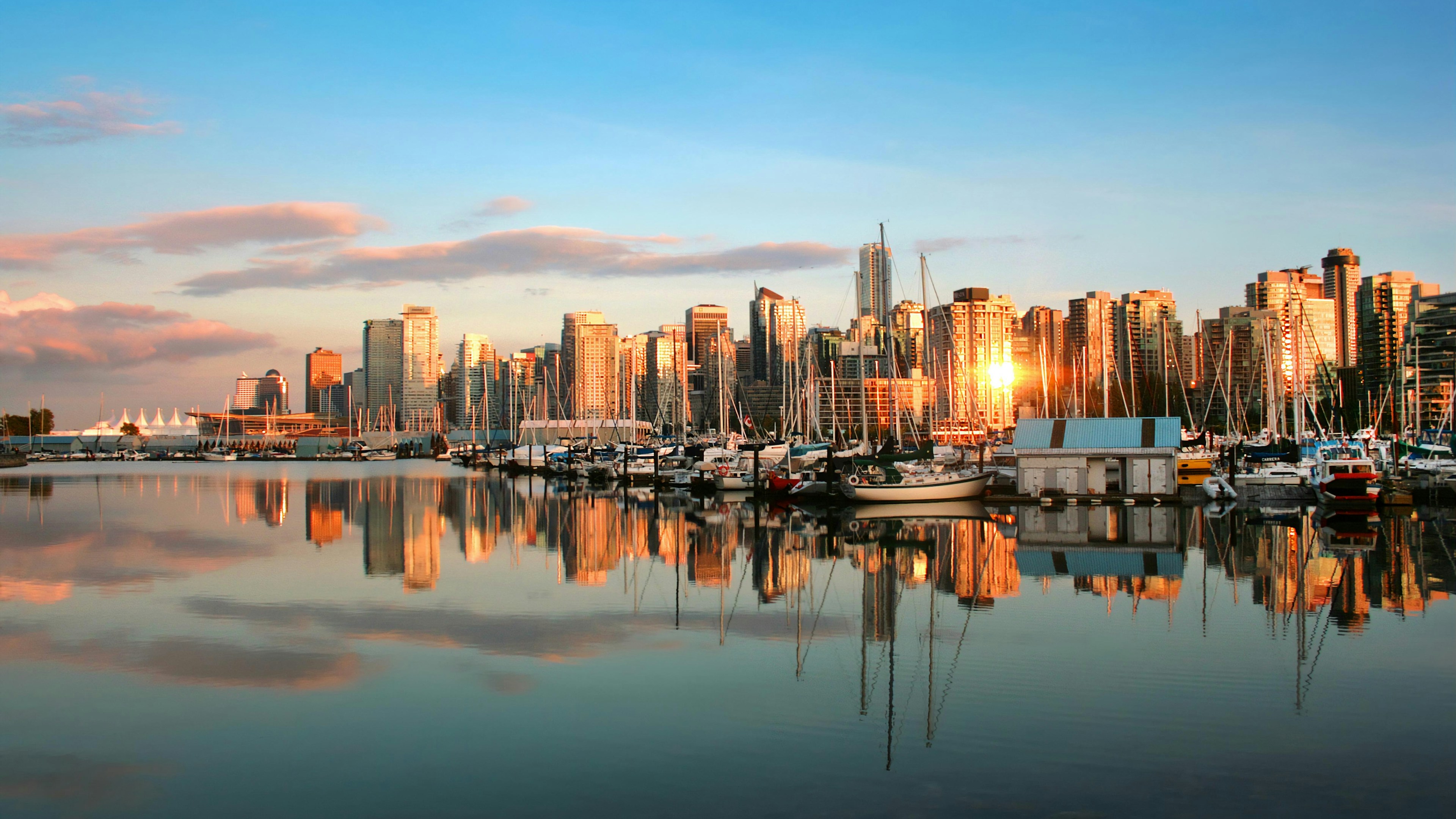 A marina where boats are moored in calm waters that reflect the city skyline at sunset, with tall buildings and clouds in the background.