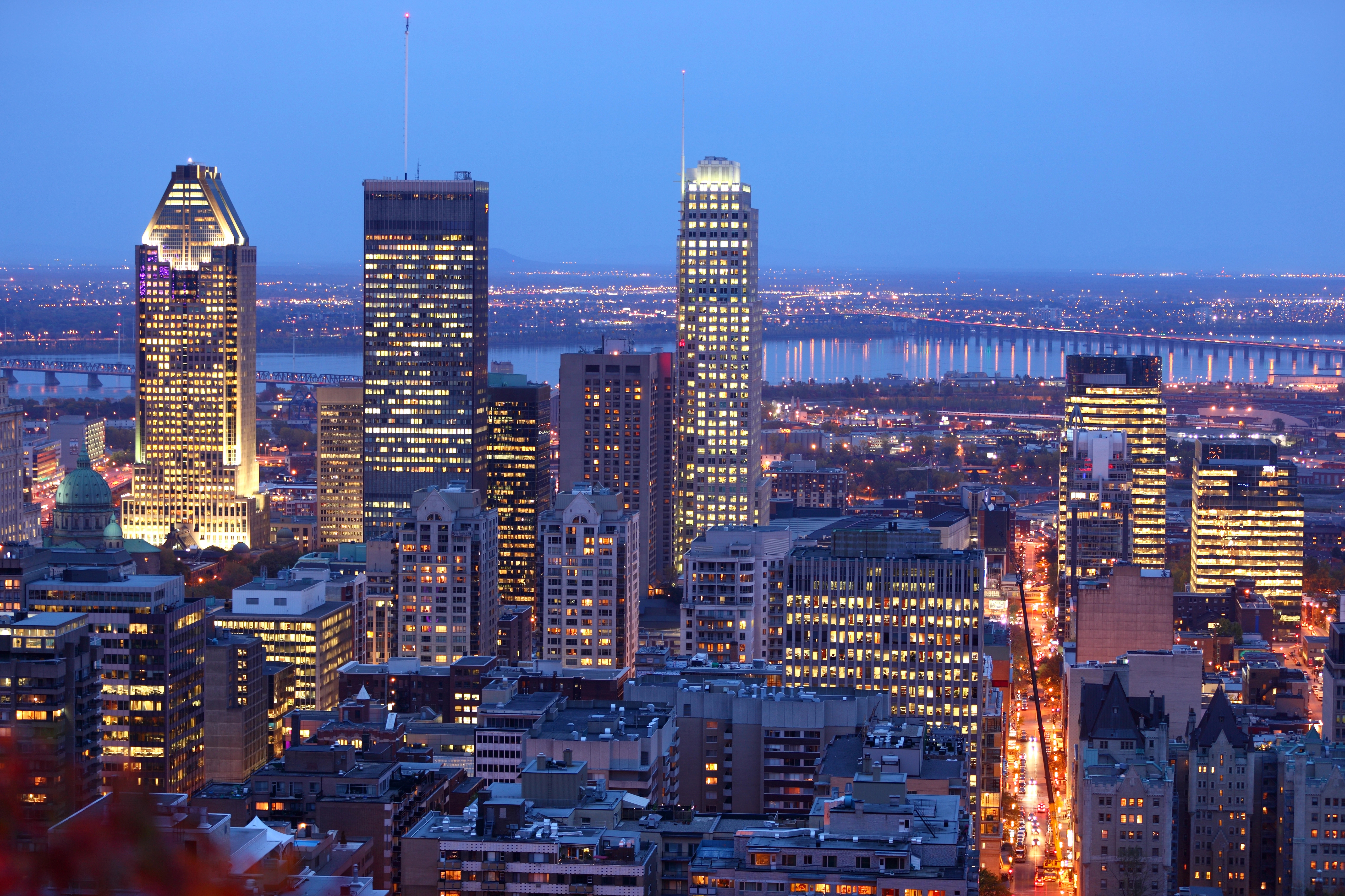 A city skyline at dusk with tall, illuminated skyscrapers and a river in the background, reflecting lights from the buildings and streets below.