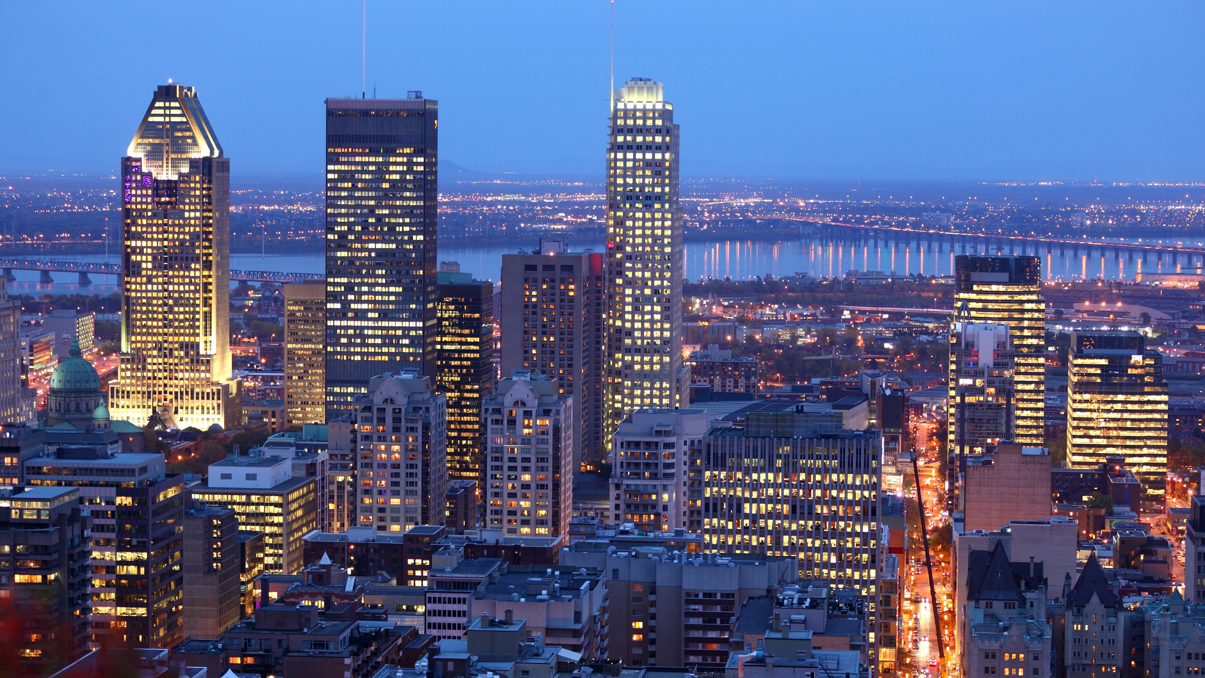 Une vue du centre-ville de Montréal au crépuscule, avec ses gratte-ciel illuminés, les lumières de la ville et le fleuve Saint-Laurent en arrière-plan, sous un ciel bleu du soir.