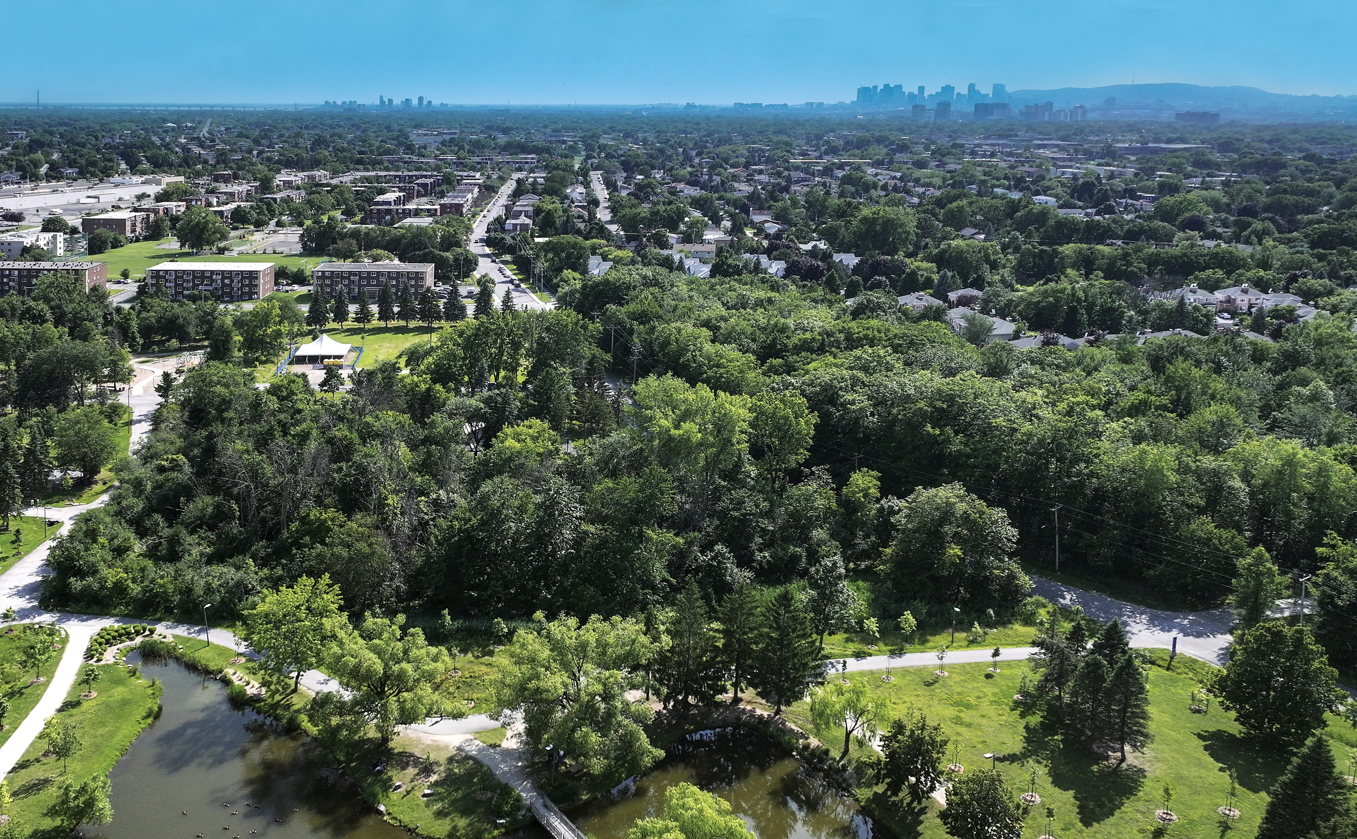 Vue aérienne d'une ville avec, au premier plan, un parc, des arbres, des sentiers pédestres, un étang, des immeubles d'habitation et, au loin, la silhouette des toits sous un ciel bleu limpide.