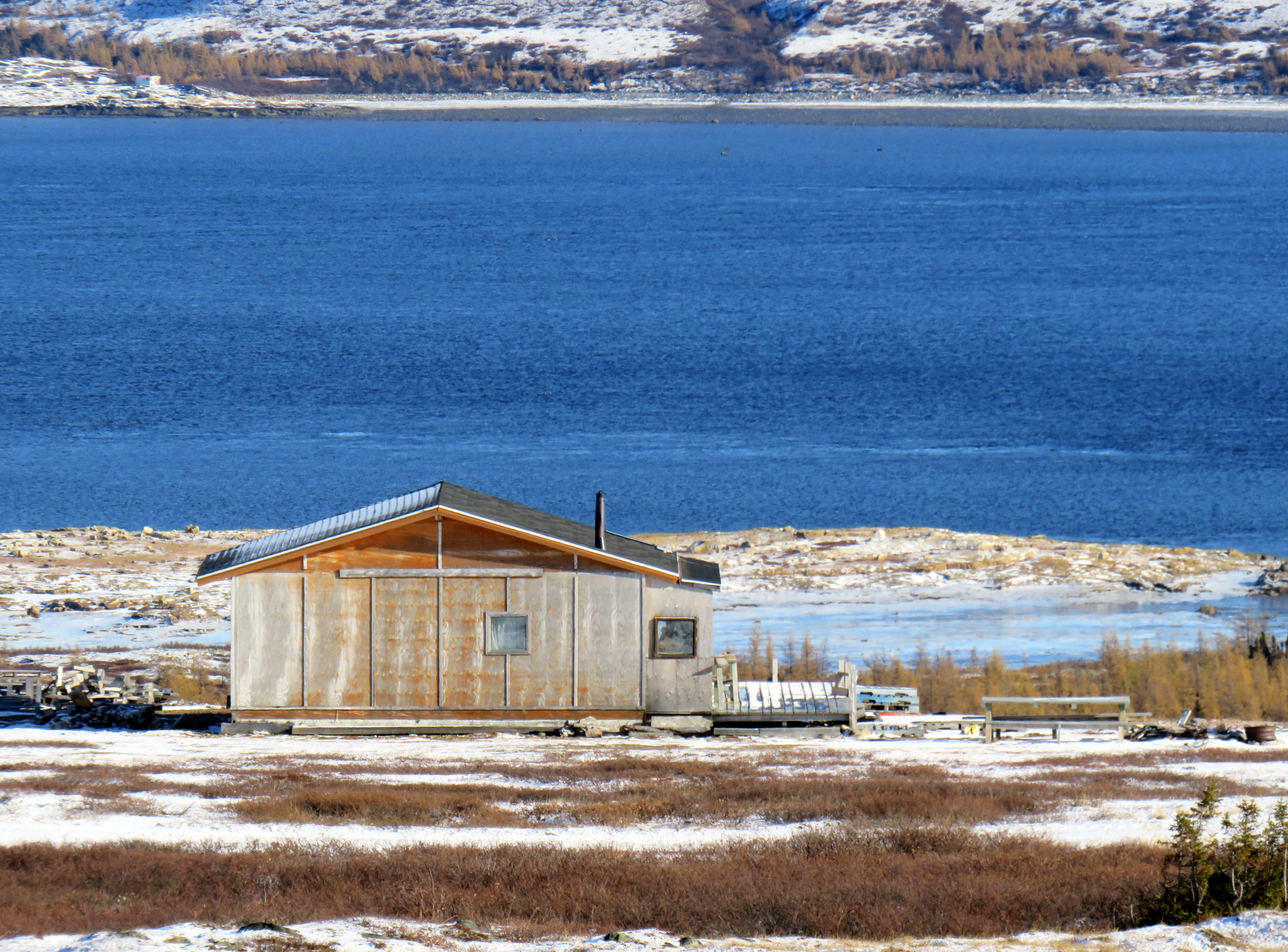 Une petite cabane en bois au toit métallique se dresse sur un sol enneigé, près d'un lac bleu, entourée d'une végétation clairsemée et de collines lointaines.