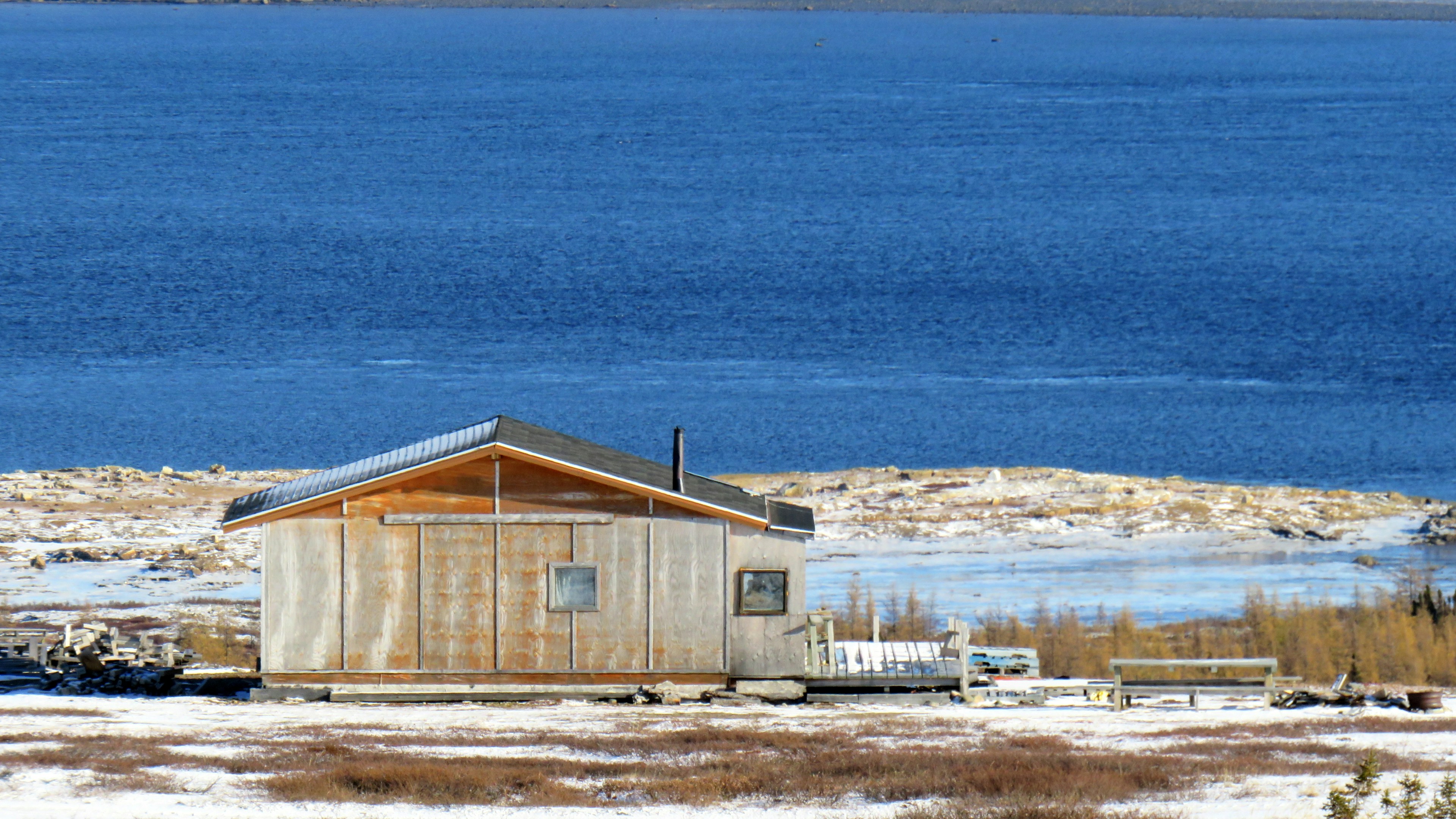 Une petite cabane en bois au toit métallique se dresse sur un sol enneigé, près d'un lac bleu, entourée d'une végétation clairsemée et de collines lointaines.