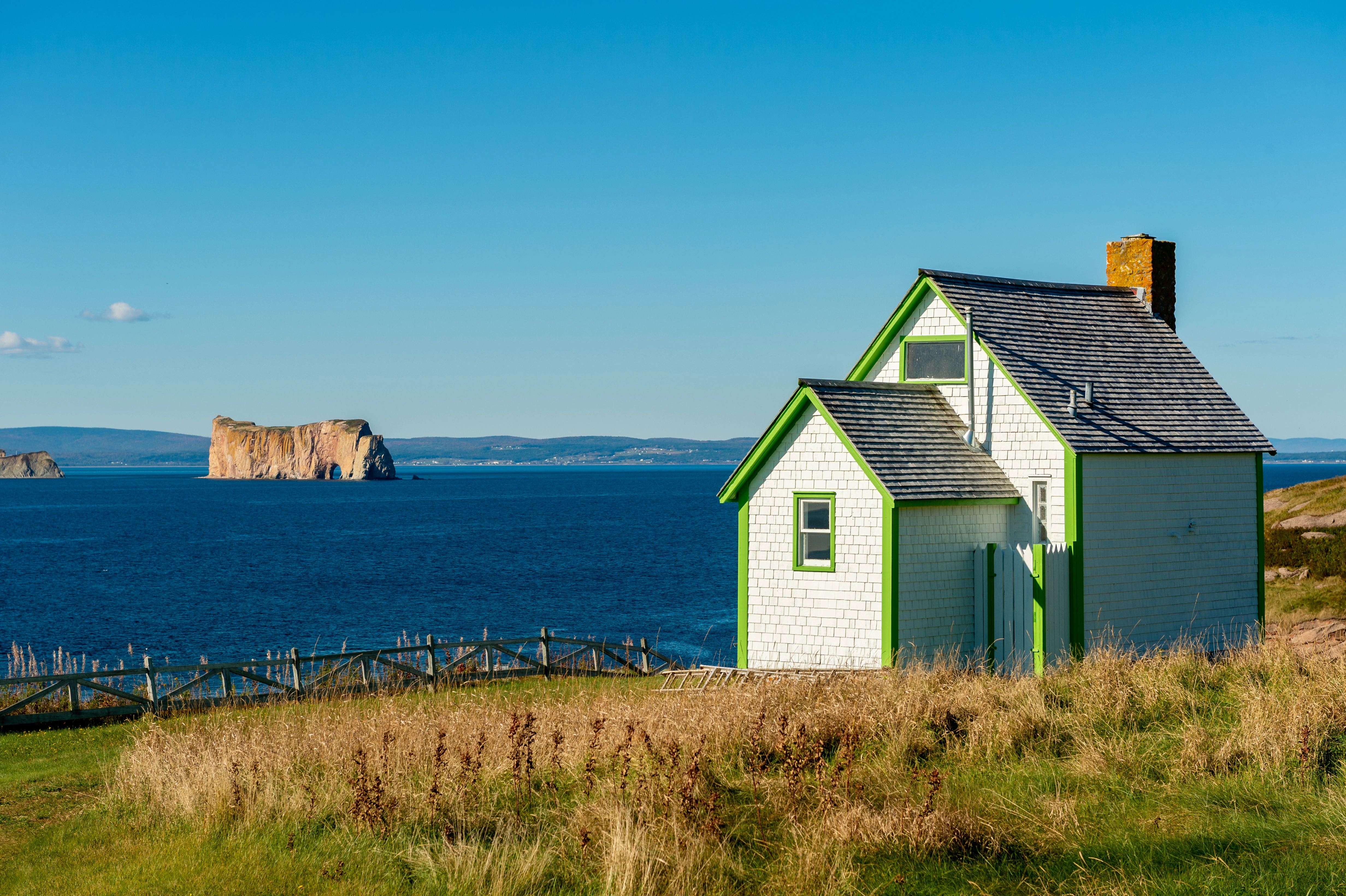 Une petite maison blanche aux moulures vertes se dresse sur une colline verdoyante surplombant la mer bleue, avec en arrière-plan un grand rocher émergeant de l'eau sous un ciel bleu limpide.