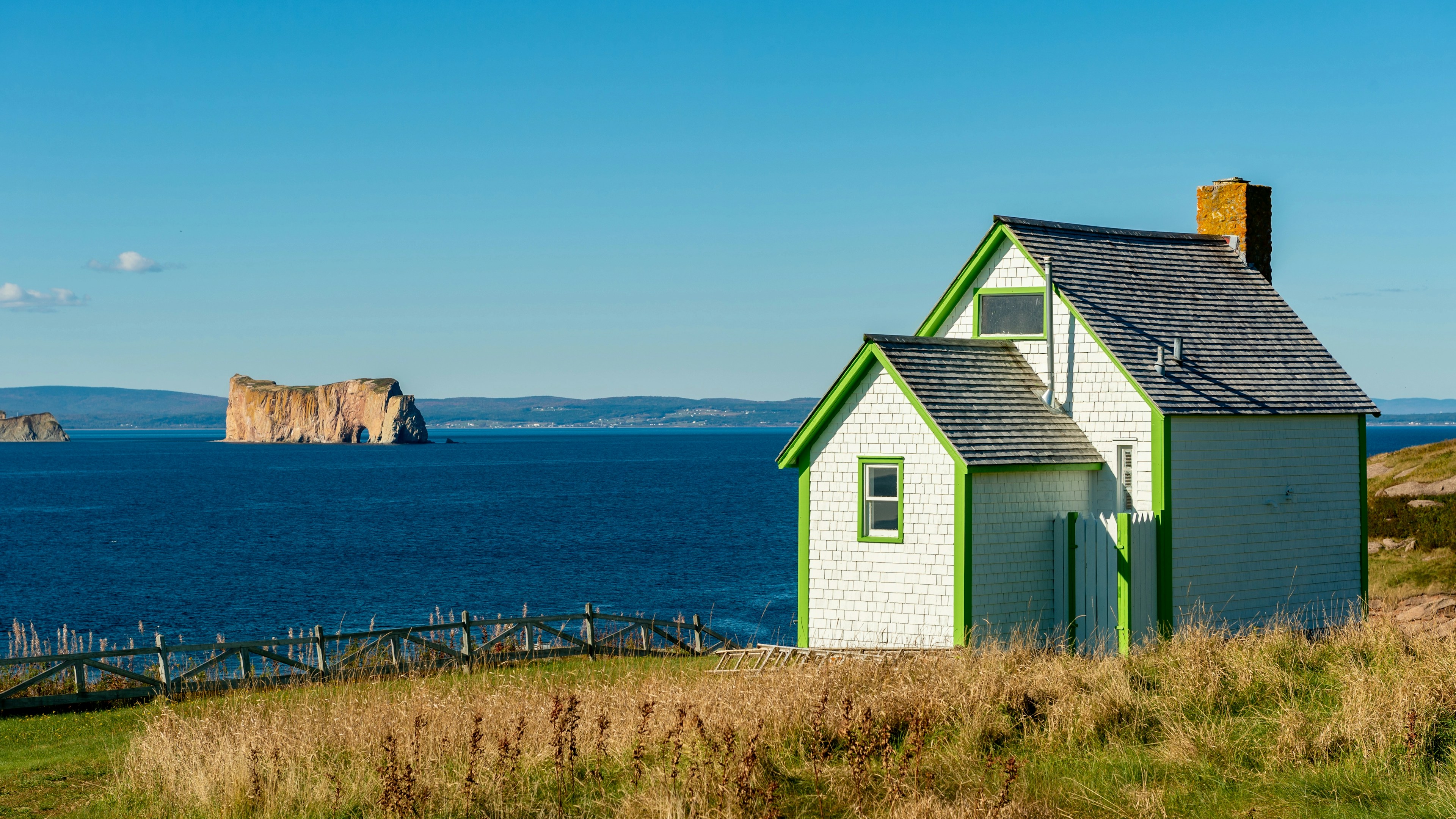 Une petite maison blanche aux moulures vertes se dresse sur une colline verdoyante surplombant la mer bleue, avec en arrière-plan un grand rocher émergeant de l'eau sous un ciel bleu limpide.