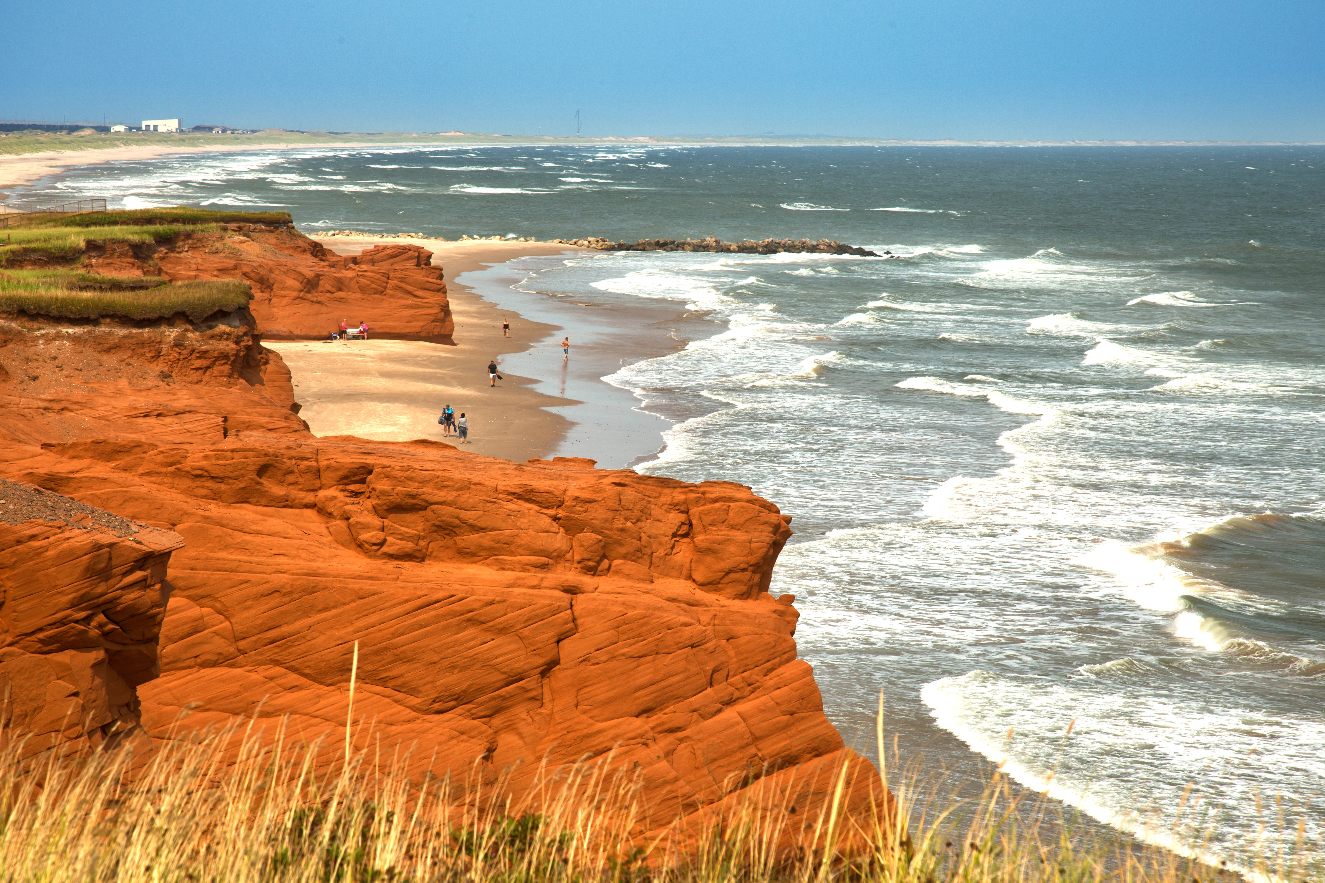 Des falaises de grès rouge surplombent une plage de sable où les vagues viennent s'écraser sur le rivage. Quelques personnes se promènent au bord de l'eau, et de l'herbe pousse au pied de la falaise, sous un ciel partiellement nuageux.