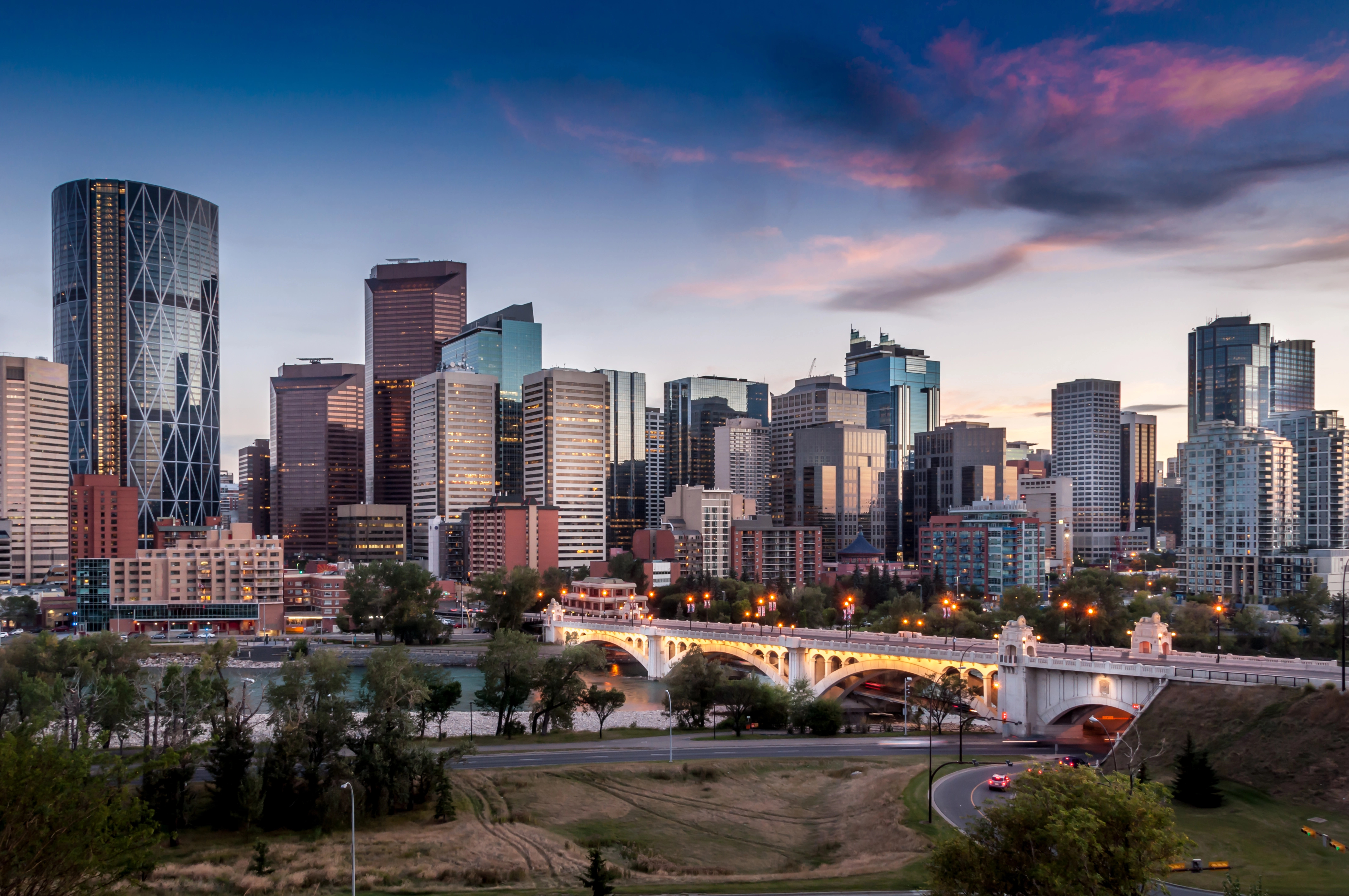 Panorama du centre-ville au crépuscule, avec ses gratte-ciel modernes, un pont illuminé enjambant une rivière et des voitures circulant sur les routes en contrebas, sous un ciel partiellement nuageux.