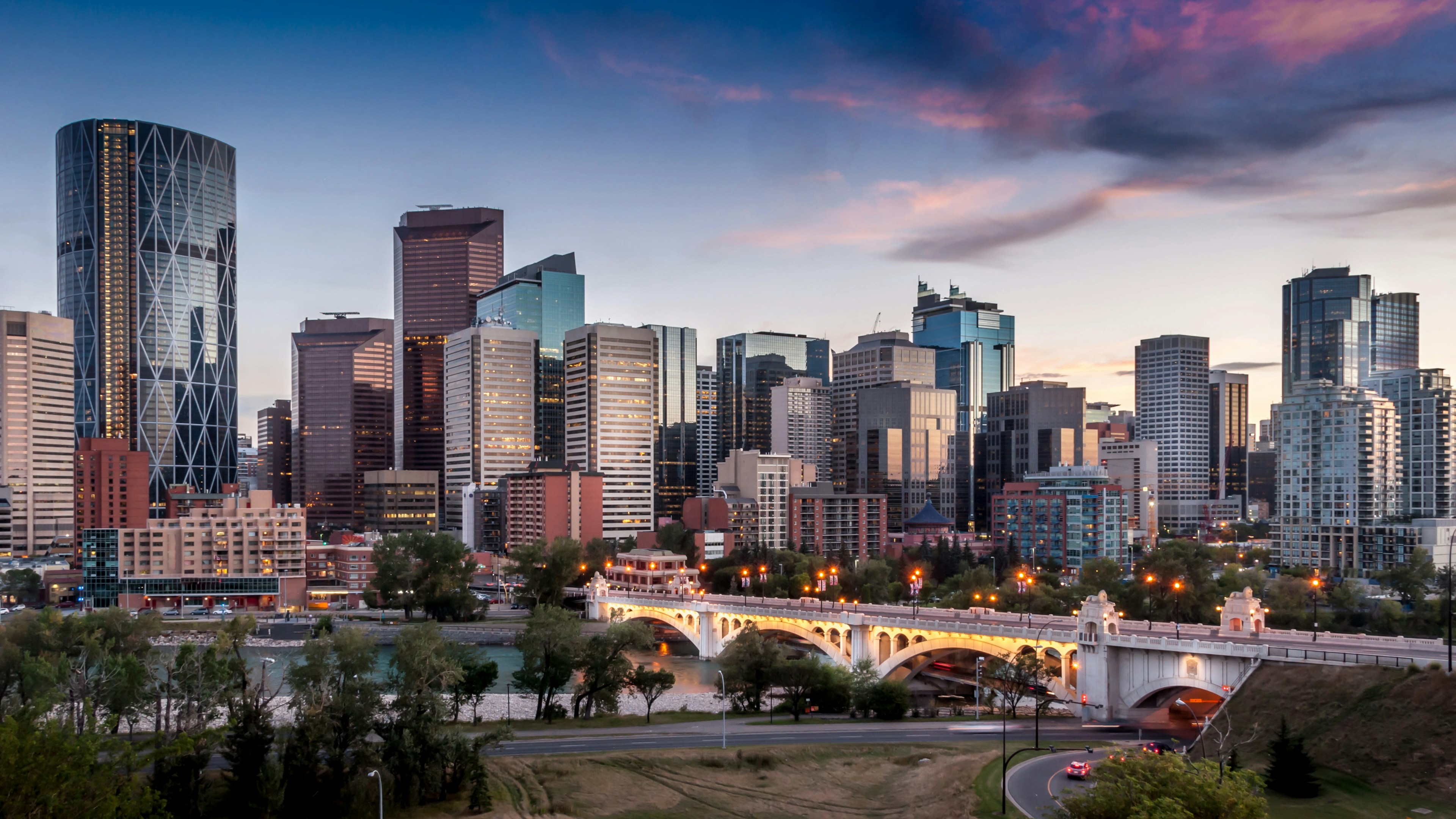 Panorama du centre-ville au crépuscule, avec ses gratte-ciel modernes, un pont illuminé enjambant une rivière et des voitures circulant sur les routes en contrebas, sous un ciel partiellement nuageux.