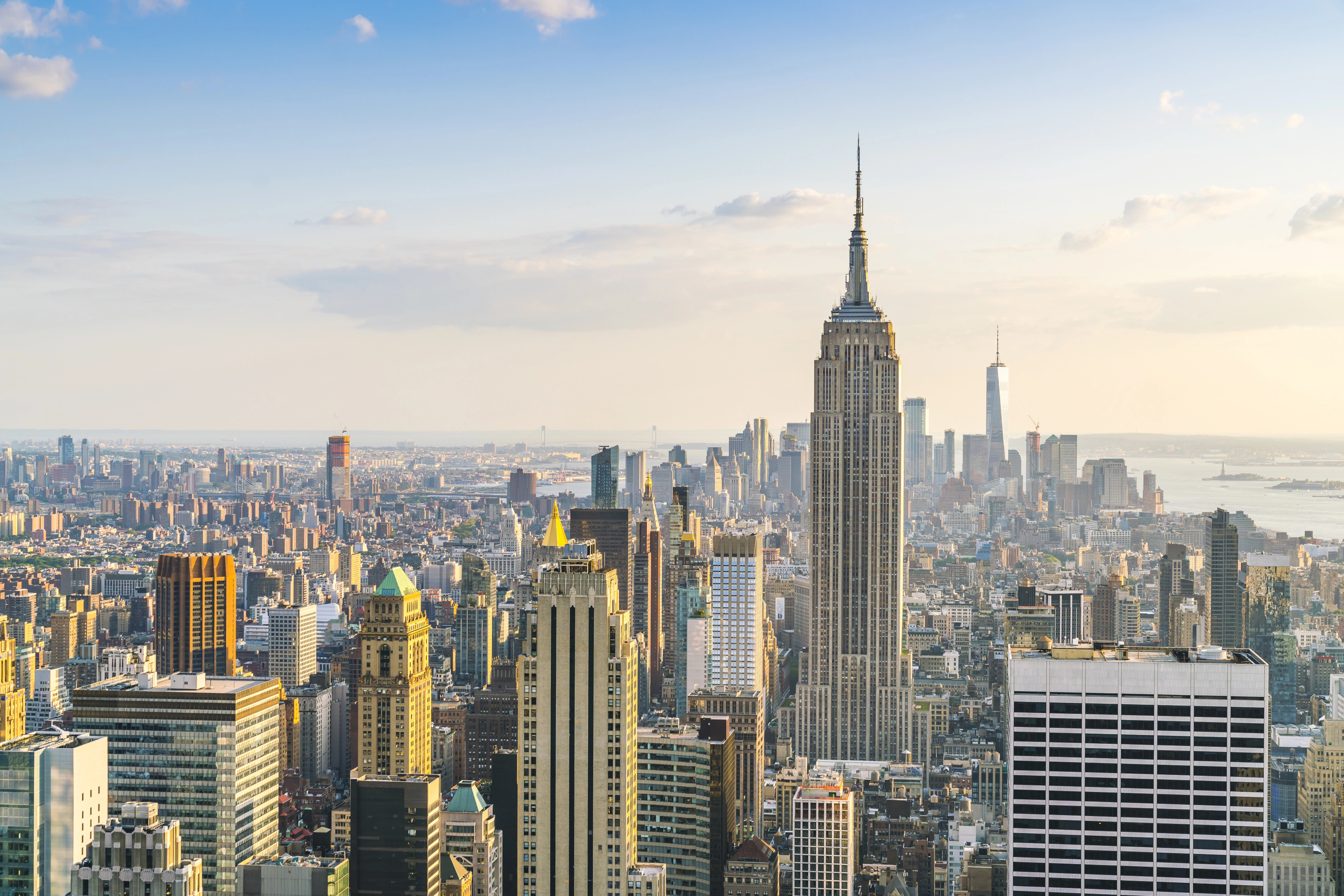 A city skyline with tall skyscrapers under a partly cloudy sky, featuring a prominent, towering building in the center. The scene is bright with distant water visible in the background.