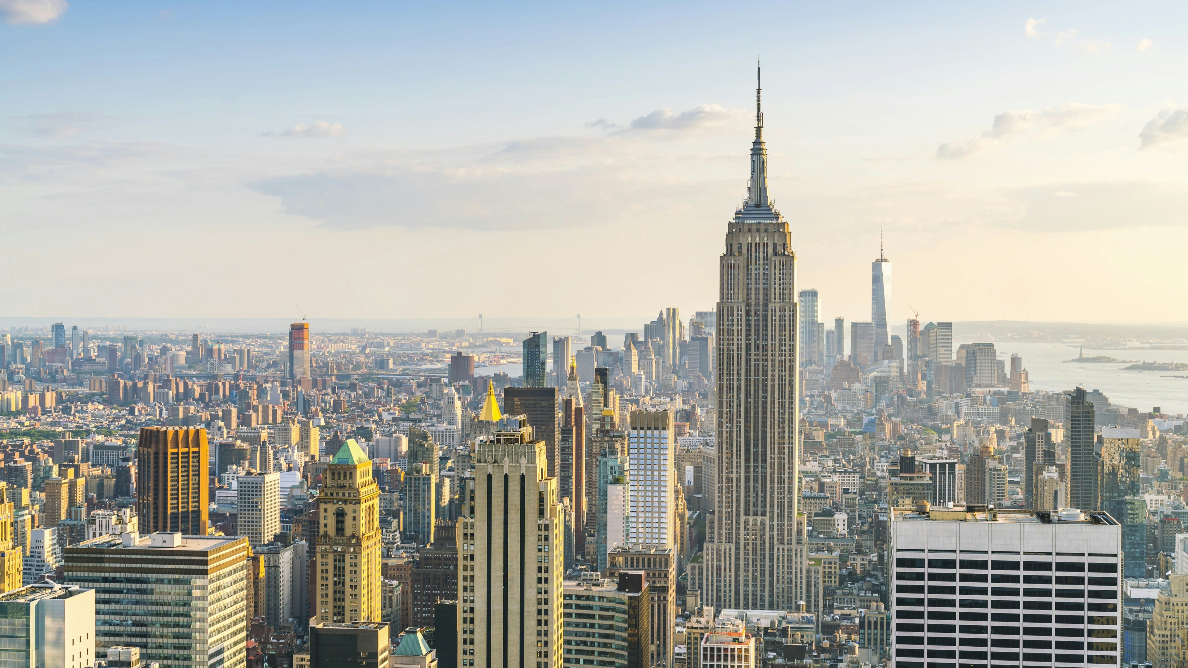 A city skyline with tall skyscrapers under a partly cloudy sky, featuring a prominent, towering building in the center. The scene is bright with distant water visible in the background.