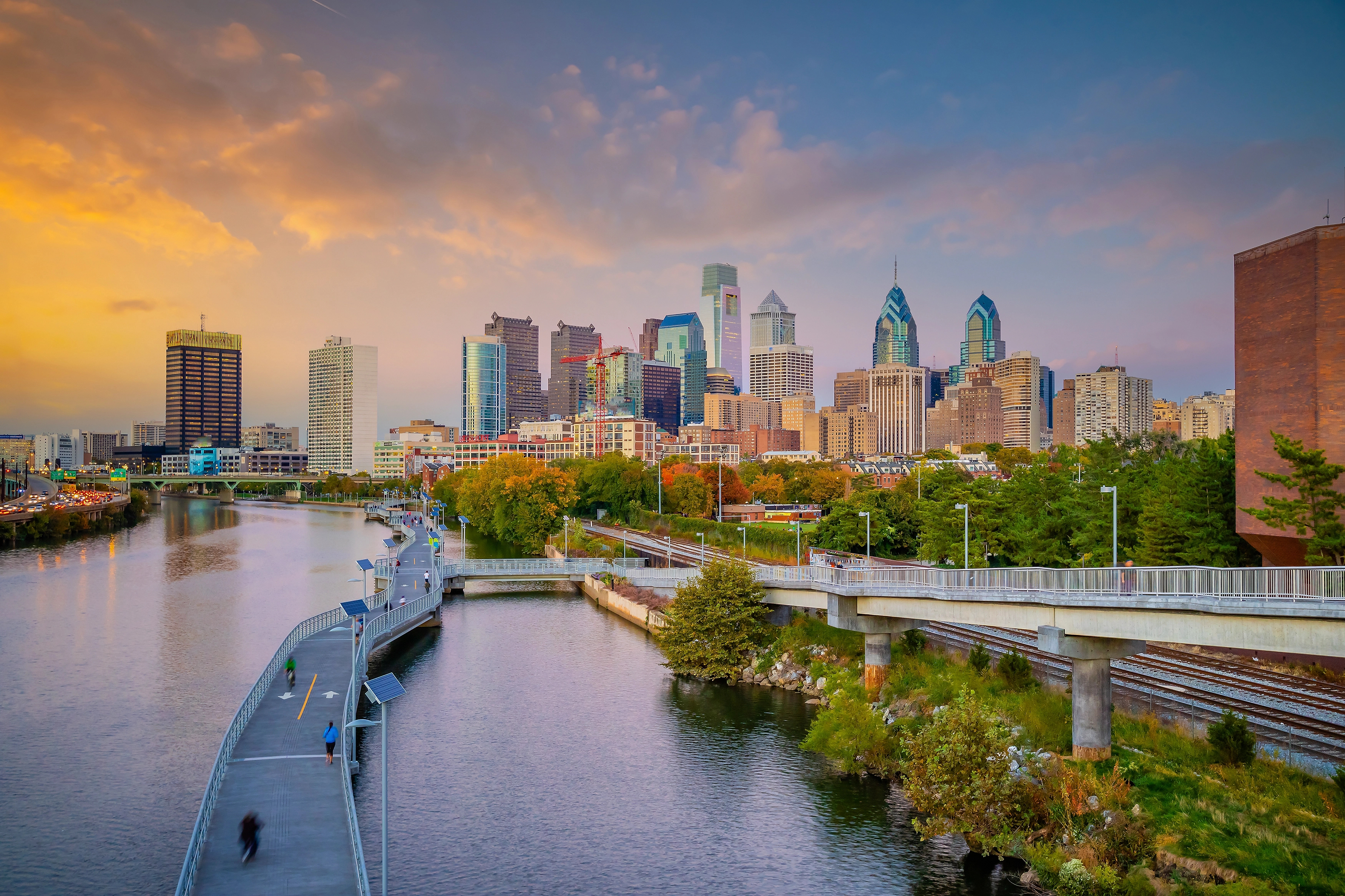 Philadelphia skyline at sunset with skyscrapers, a river, a walking path, and a bridge in the foreground surrounded by greenery.