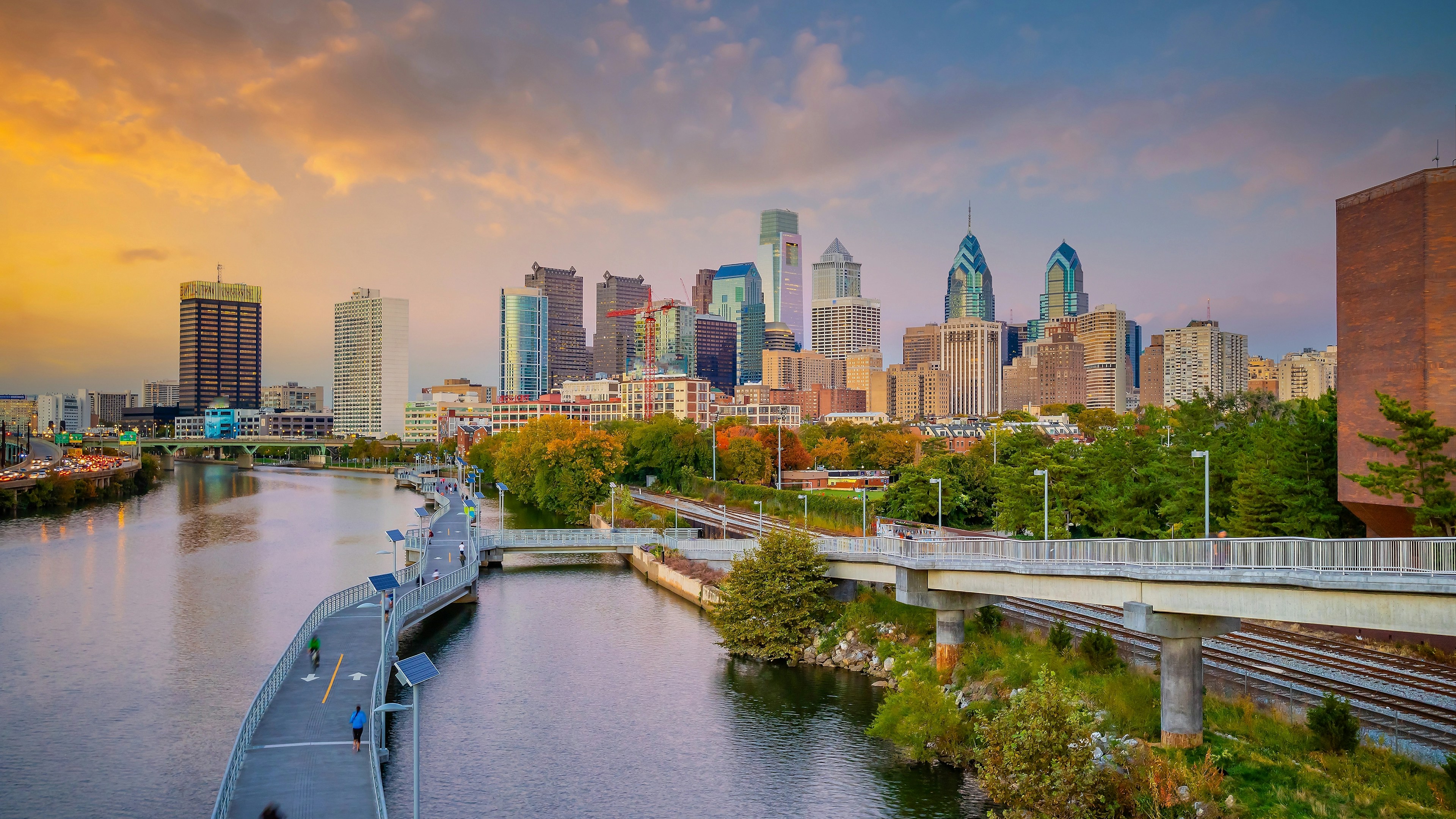 Philadelphia skyline at sunset with skyscrapers, a river, a walking path, and a bridge in the foreground surrounded by greenery.