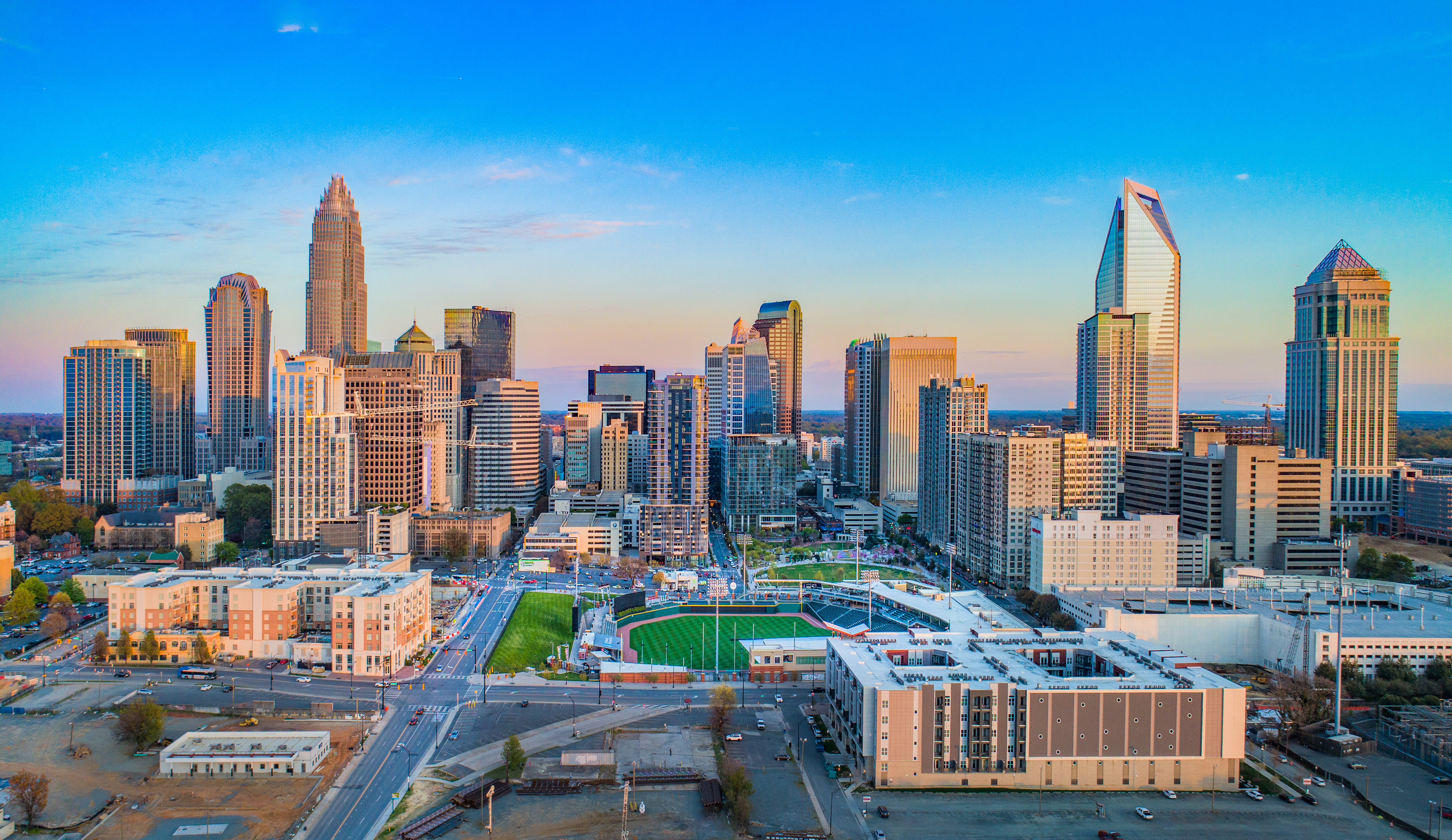 Aerial view of a city skyline at sunset, featuring tall modern skyscrapers, a sports field, and various buildings under a clear blue sky.