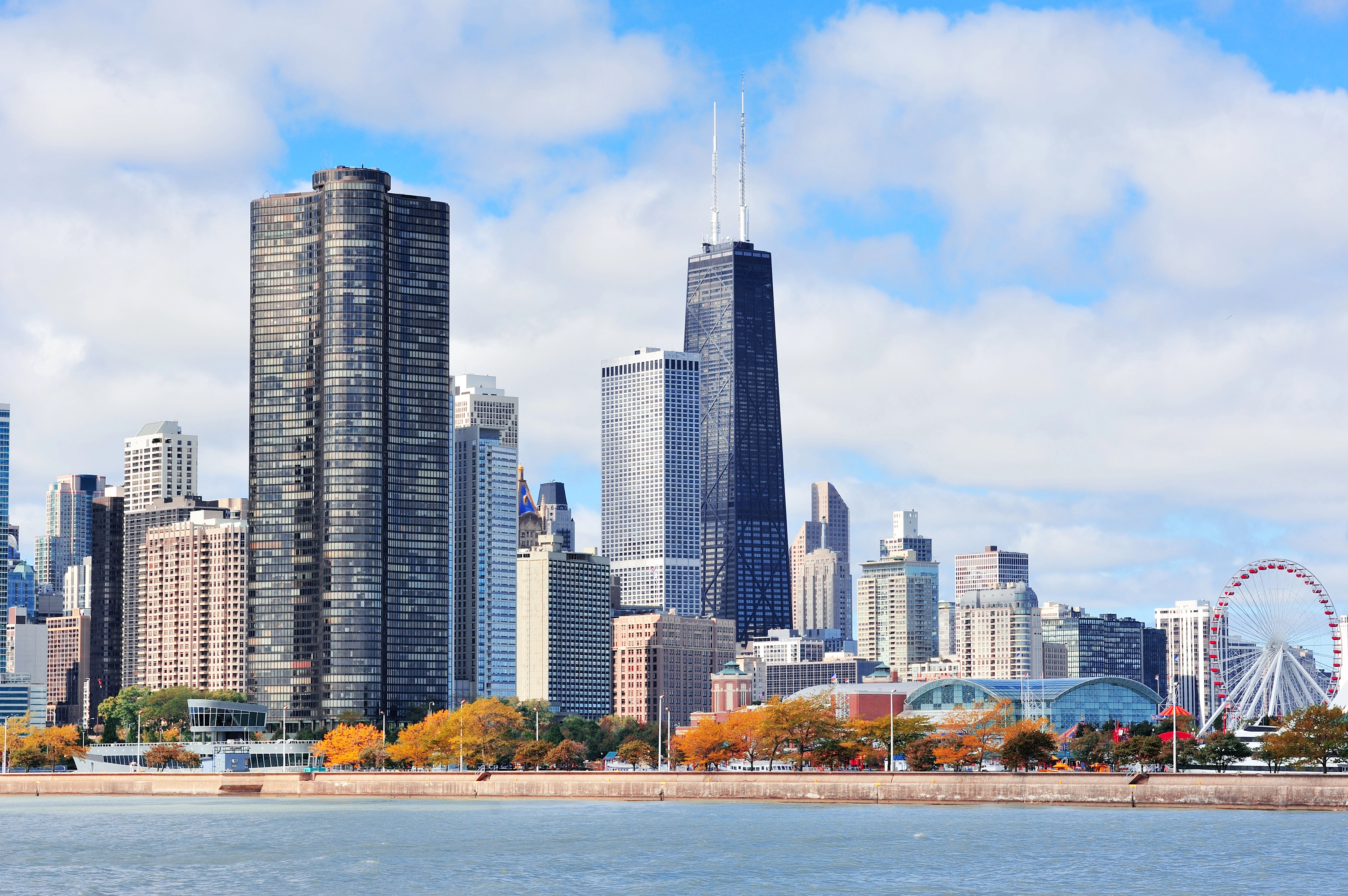 La ligne d'horizon de Chicago, avec ses grands gratte-ciel, une grande roue près du bord de l'eau et des arbres aux feuilles d'automne sous un ciel partiellement nuageux. Le lac Michigan est visible au premier plan.