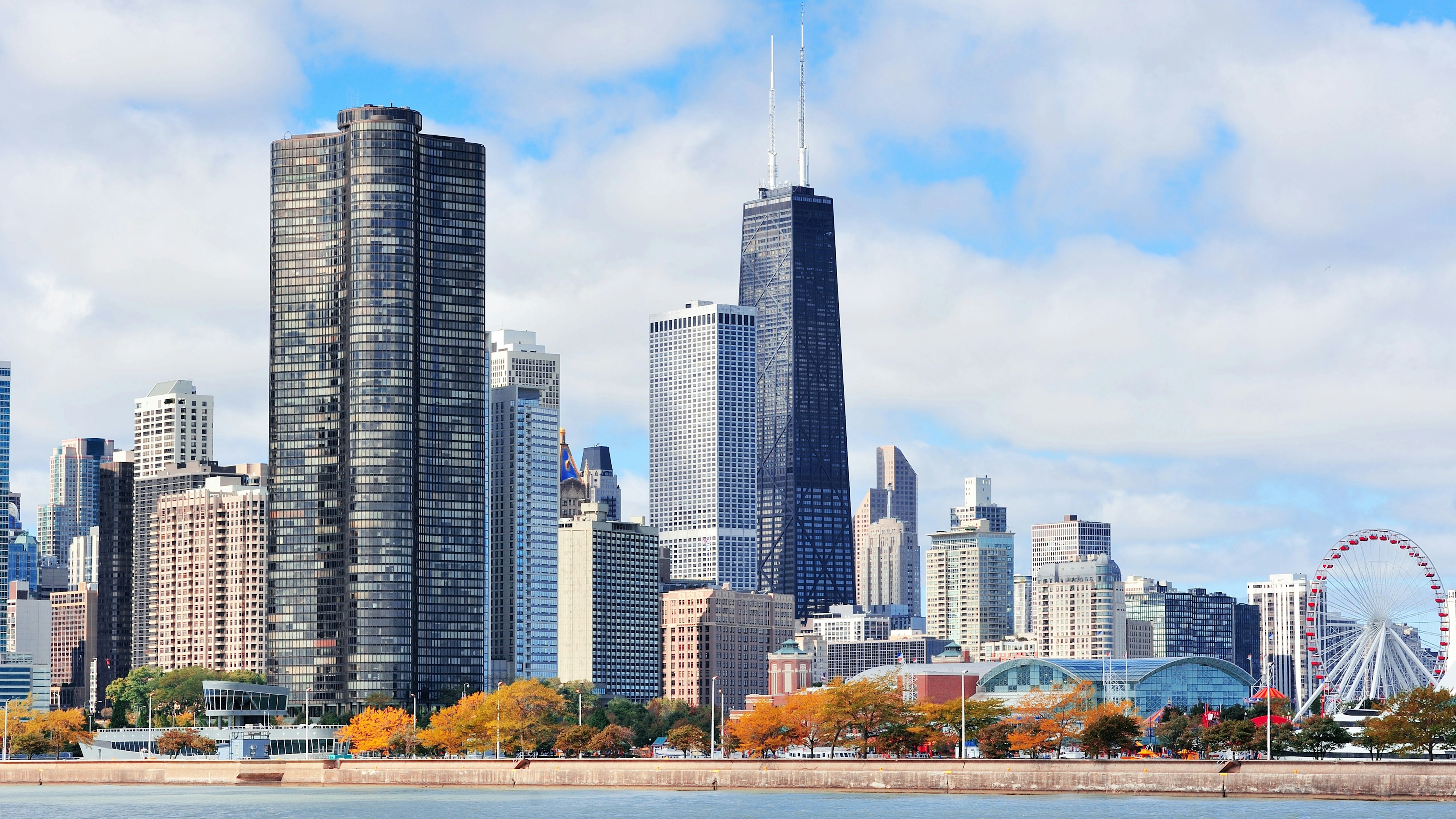 La ligne d'horizon de Chicago, avec ses grands gratte-ciel, une grande roue près du bord de l'eau et des arbres aux feuilles d'automne sous un ciel partiellement nuageux. Le lac Michigan est visible au premier plan.