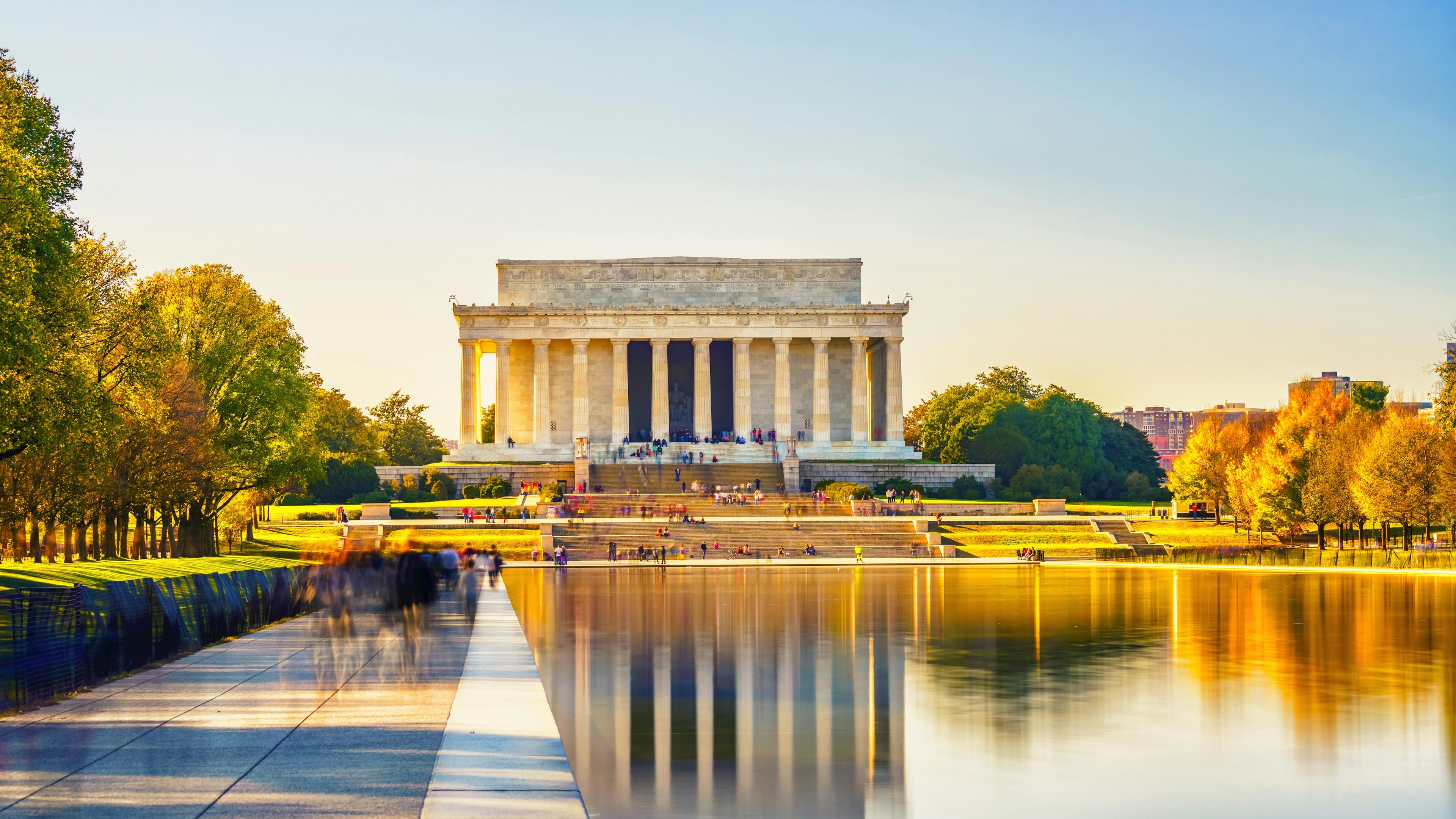 Le Lincoln Memorial, à Washington, D.C., se dresse au bout du Reflecting Pool, entouré d'arbres et de passants, sous un ciel bleu limpide baigné par la lumière dorée du soleil.