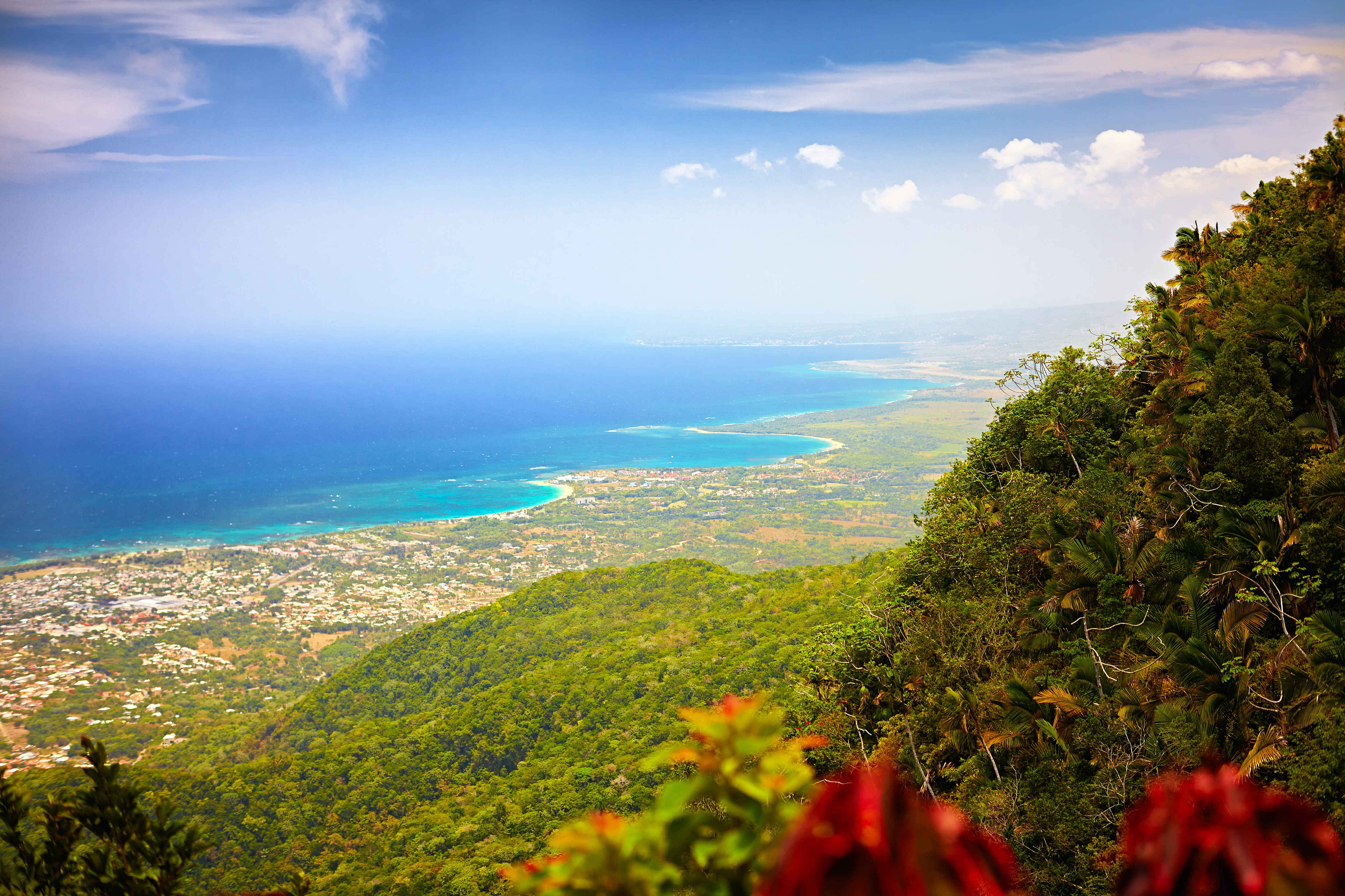 Une colline verdoyante surplombe une ville côtière aux eaux turquoise et aux plages de sable blanc, sous un ciel bleu partiellement nuageux.