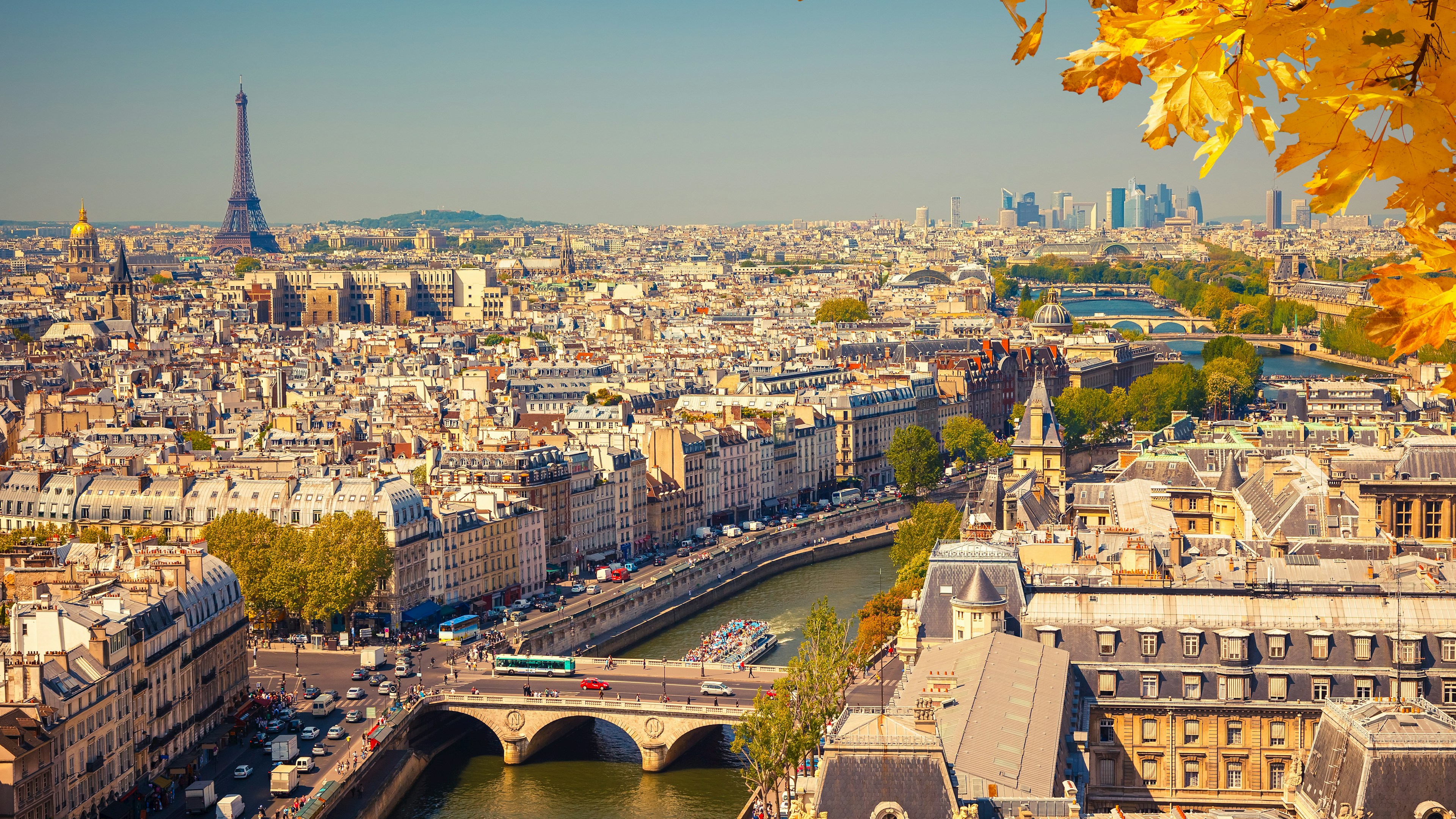 Vue aérienne de Paris avec la tour Eiffel, la Seine et des bâtiments historiques sous un ciel dégagé. Des feuilles d'automne jaunes encadrent le coin supérieur droit.