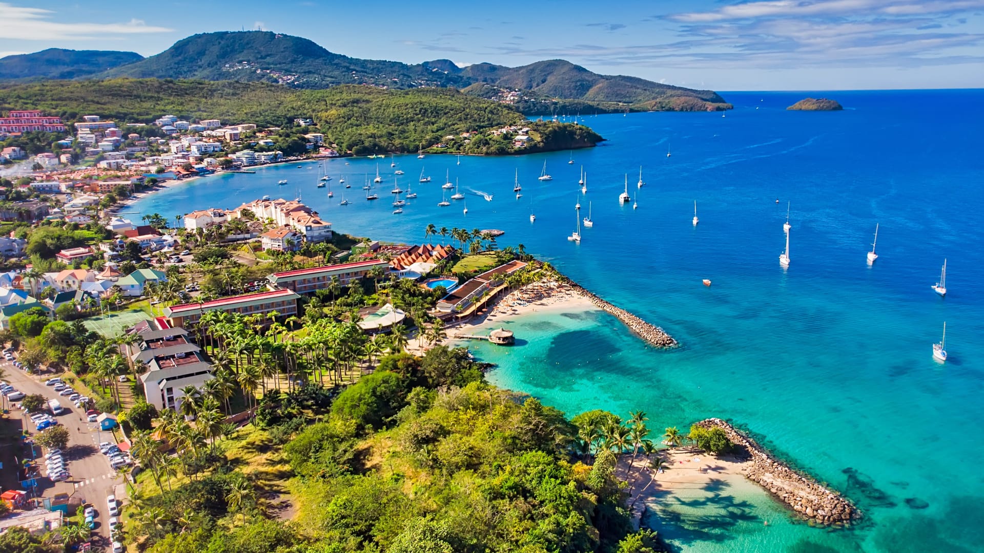An aerial view of a tropical coastal town with turquoise waters, boats moored in the bay, sandy beaches, lush vegetation, and hills in the background, all under a bright blue sky.