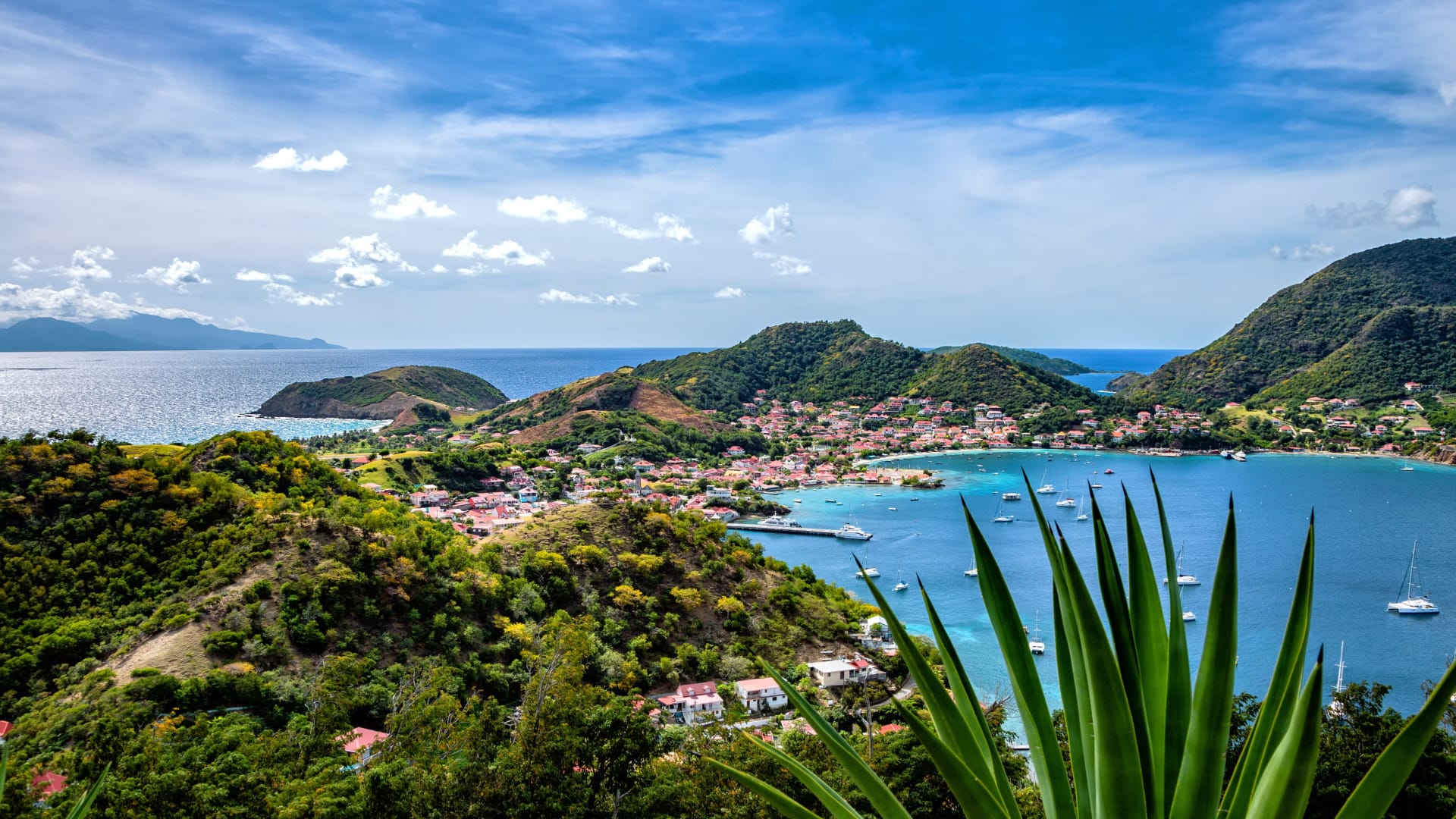 Vue d'un village côtier entouré de collines verdoyantes, d'une mer turquoise, de bateaux amarrés dans la baie et de plantes tropicales au premier plan, sous un ciel partiellement nuageux.